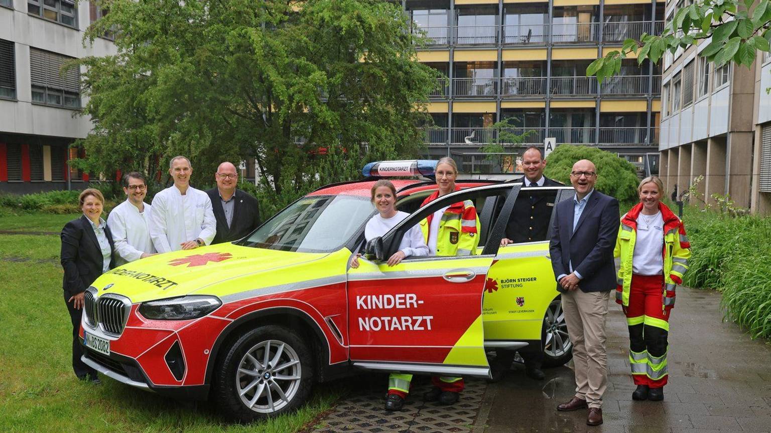 Ein Notarztwagen in Rot und Neongelb, auf der Tür steht Kindernotarzt, drei Personen in Rettungssanitäter-Kleidung und zwei Ärzte in Weiß, weitere drei Personen aus der Verwaltung und ein Mann von der Feuerwehr
