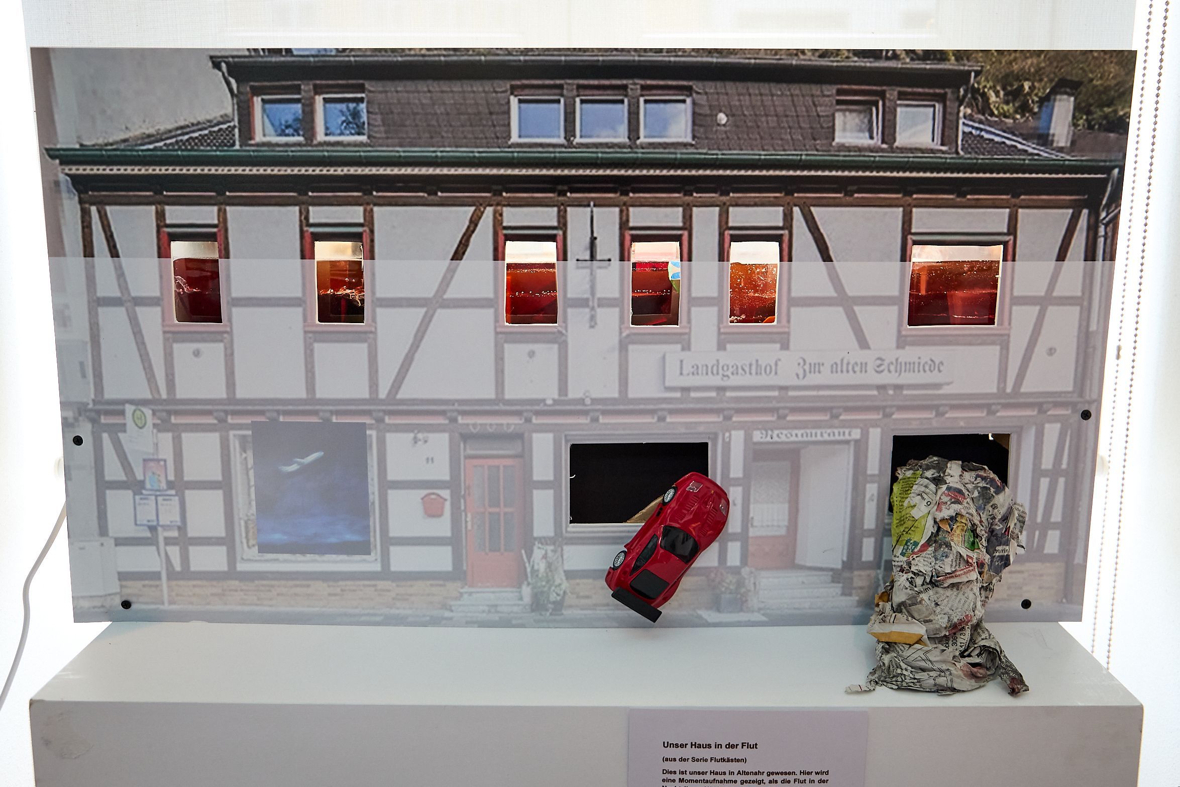 Eine matte Fläche zeigt, dass das Wasser im Fachwerkgebäude Des Landgasthofs Zur alten Schmiede bis zur Mitte des ersten Obergeschosses stand. Ein rotes Auto wurde gegen ein Fenster gespült. Aus einem anderen Fenster quillt Unrat.