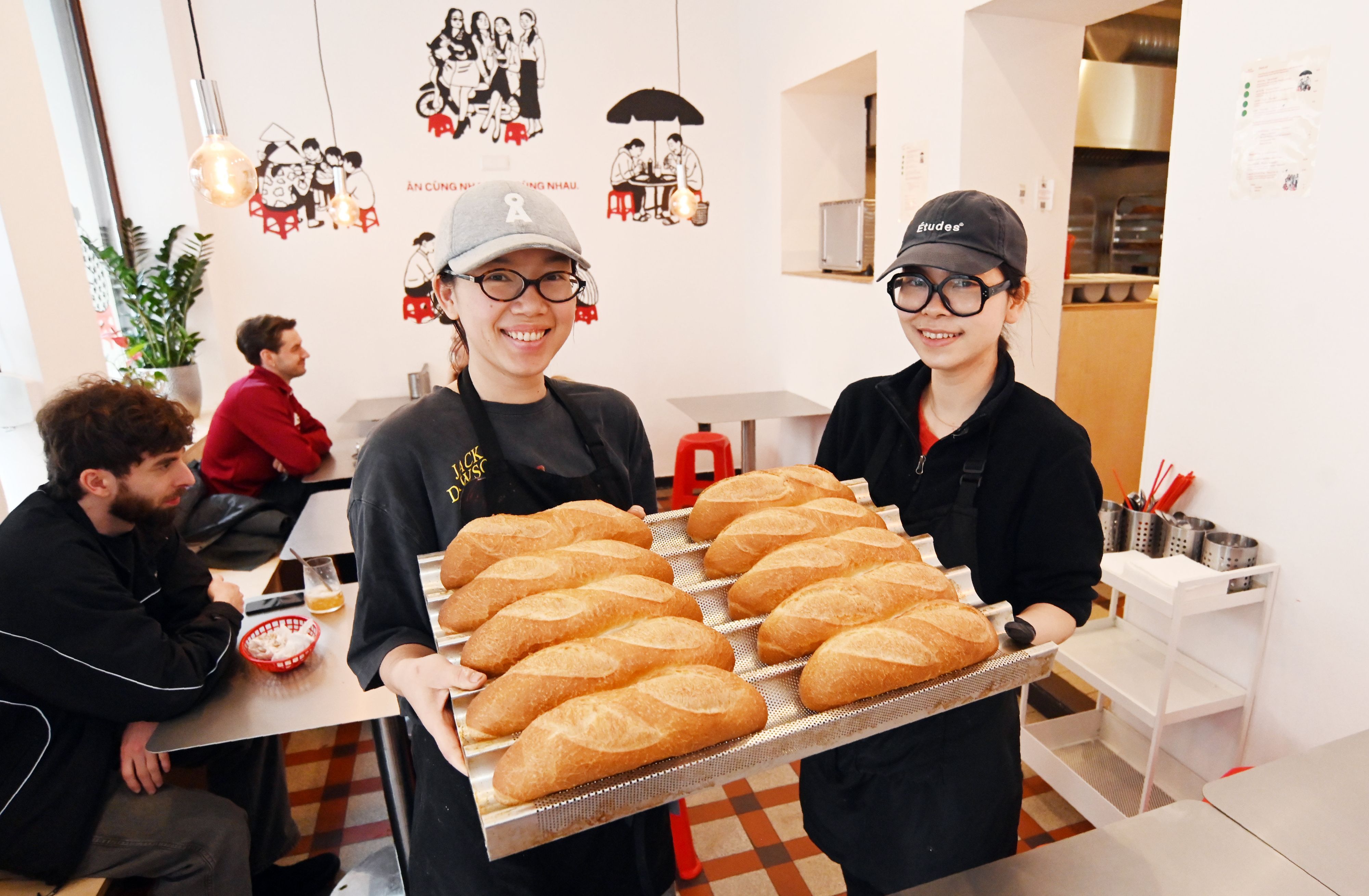 Die beiden Frauen zeigen auf einem großen Tablett die Baguettbrötchen. Beide tragen Baseball Caps und lachen in die Kamera.