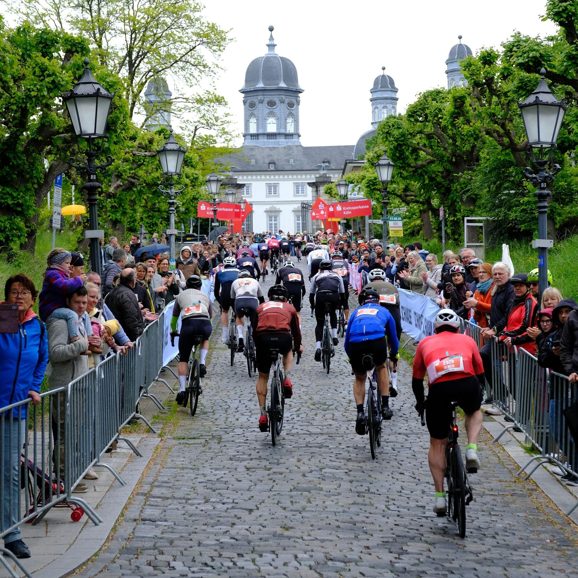 Radrennfahrer kämpfen sich über das Kopfsteinpflaster zum Bensberger Schloss hinauf. Am Rand stehen Zuschauer.