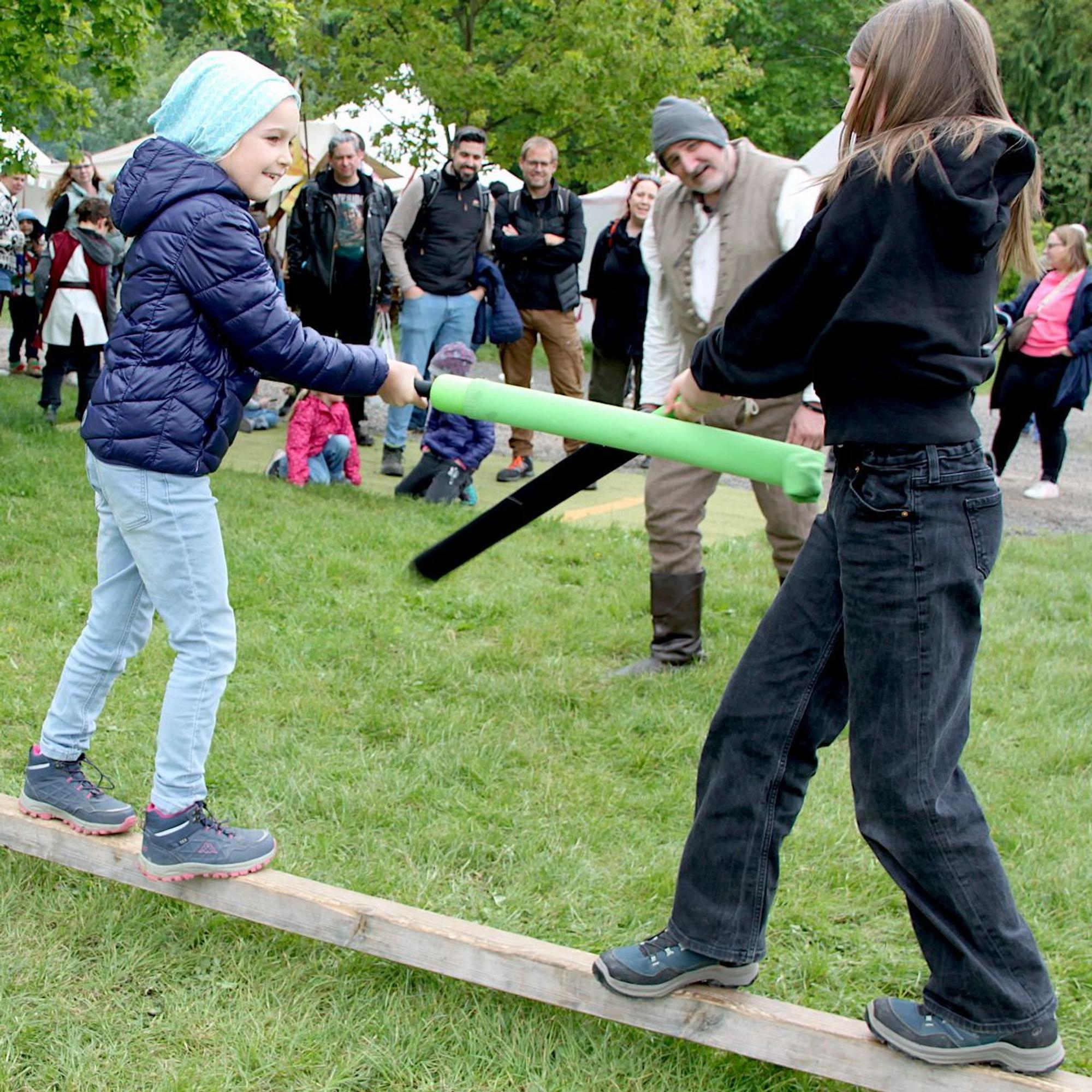 Die beiden Mädchen balancieren auf einem Balken und kämpfen mit Schaumstoff-Schwertern gegeneinander.