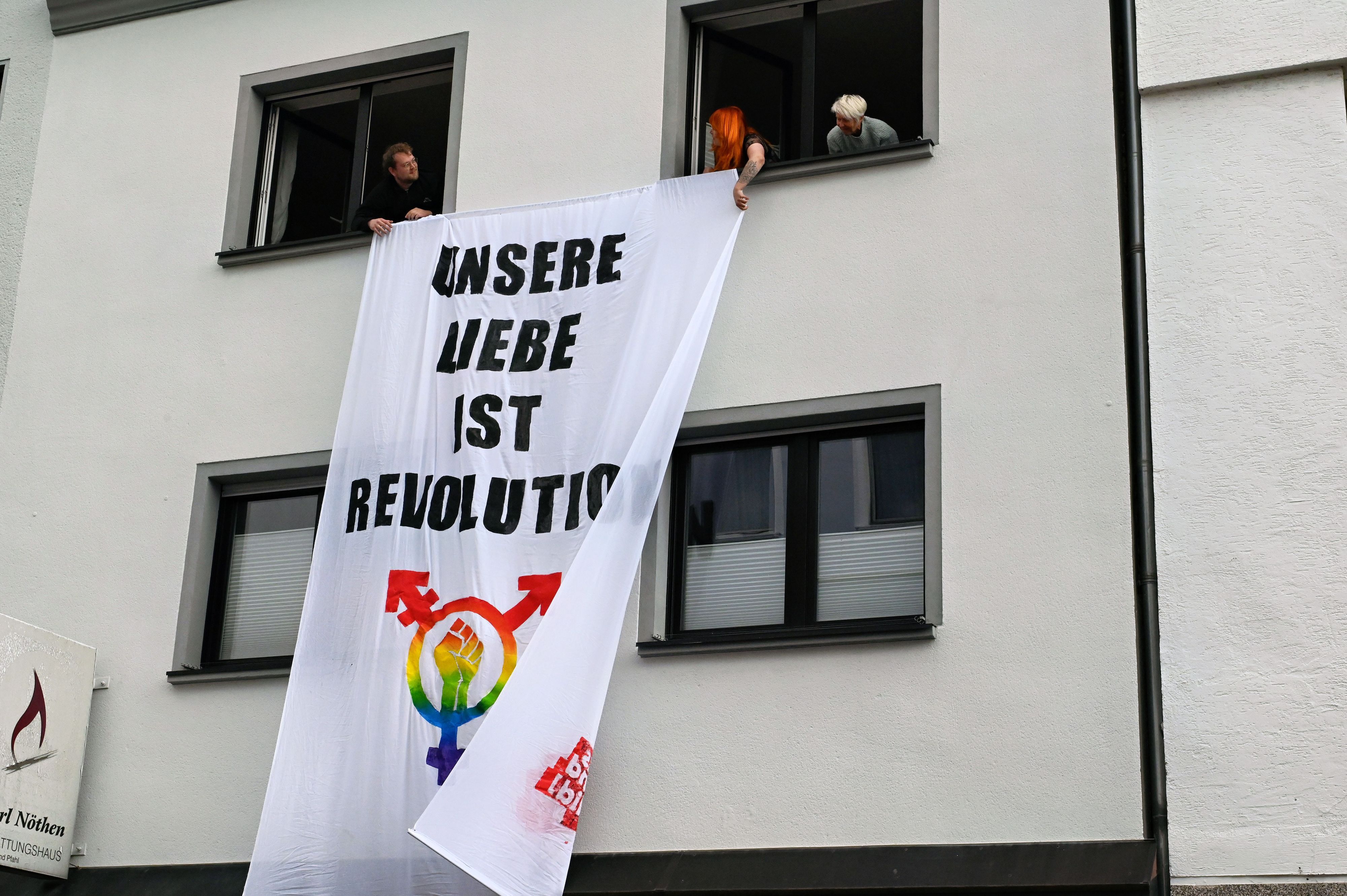 Hausbewohner hängen aus Fenstern im Obergeschoss eine Fahne mit der Aufschrift „Unsere Liebe ist Revolution“.