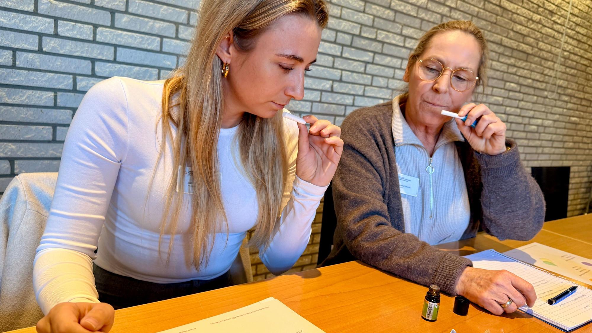 Zwei Frauen halten sich schnuppernd kleine Papierstreifen vor die Nase. Auf dem Tisch steht ein aufgeschraubtes Glasfläschchen.