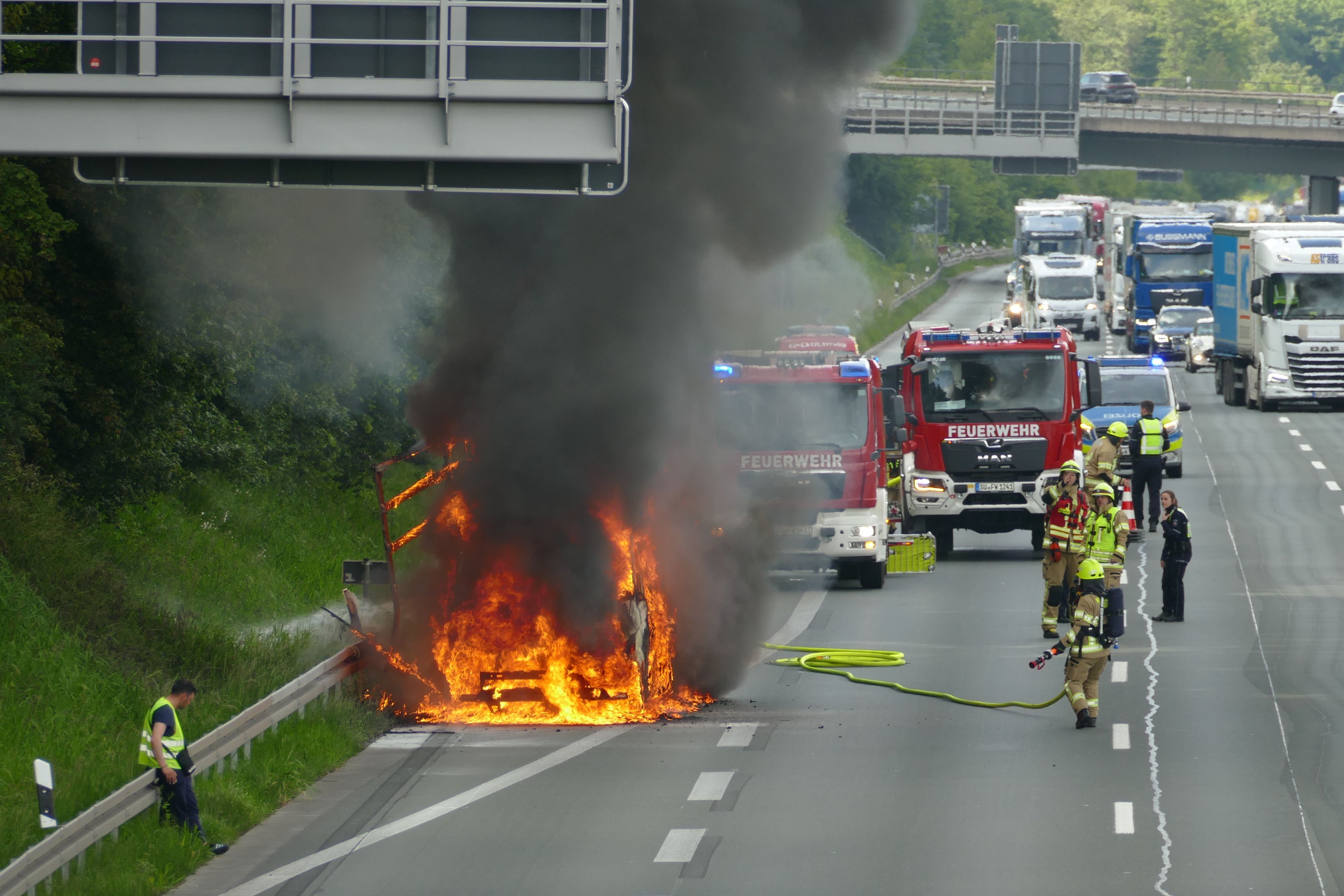 Die Aufnahme zeigt das brennende Fahrzeug und Feuerwehrleute, die mit den Löscharbeiten beginnen.