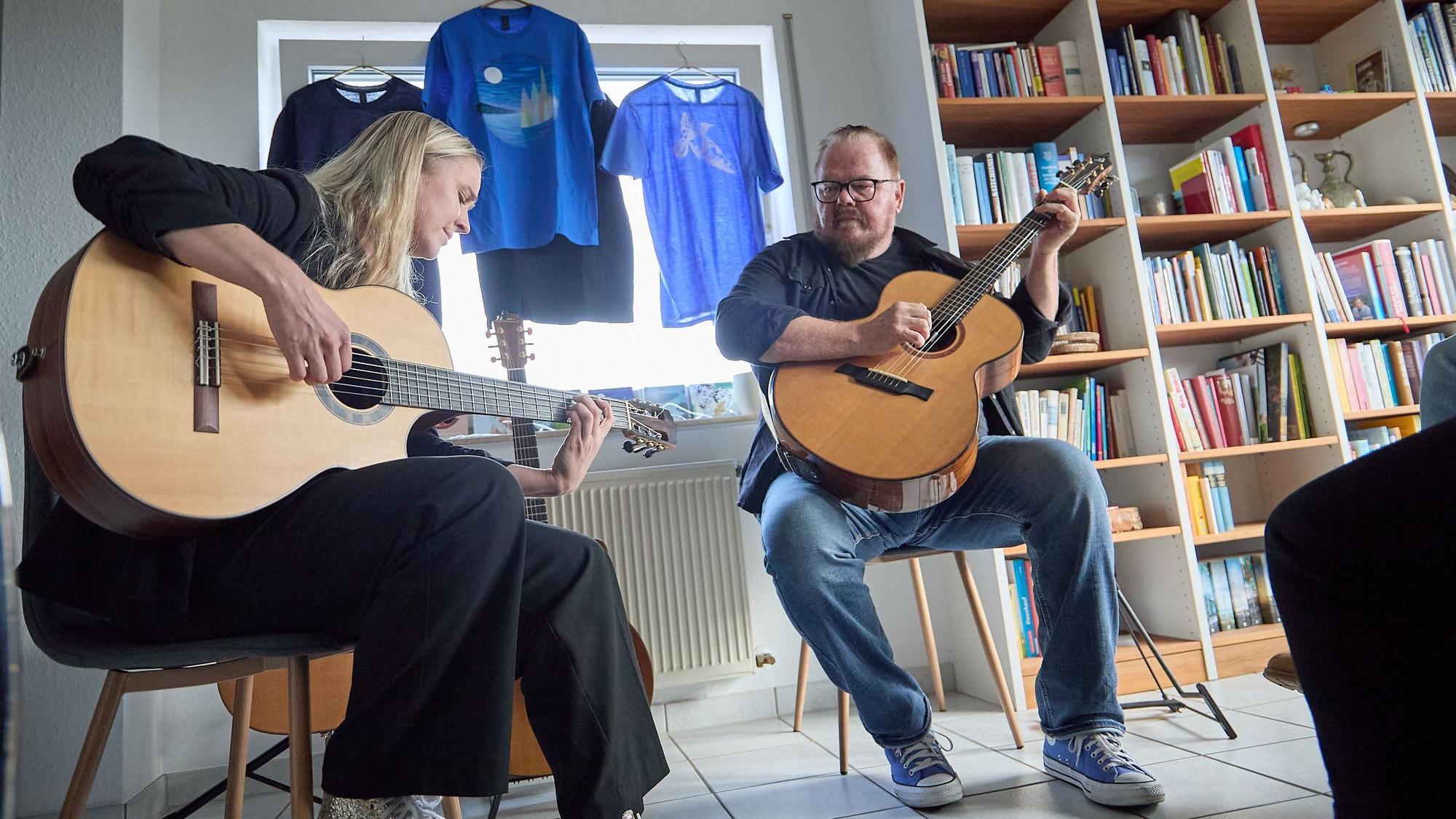 Ein Mann und eine Frau sitzen vor einem Bücherregal auf Stühlen und spielen auf Gitarren.