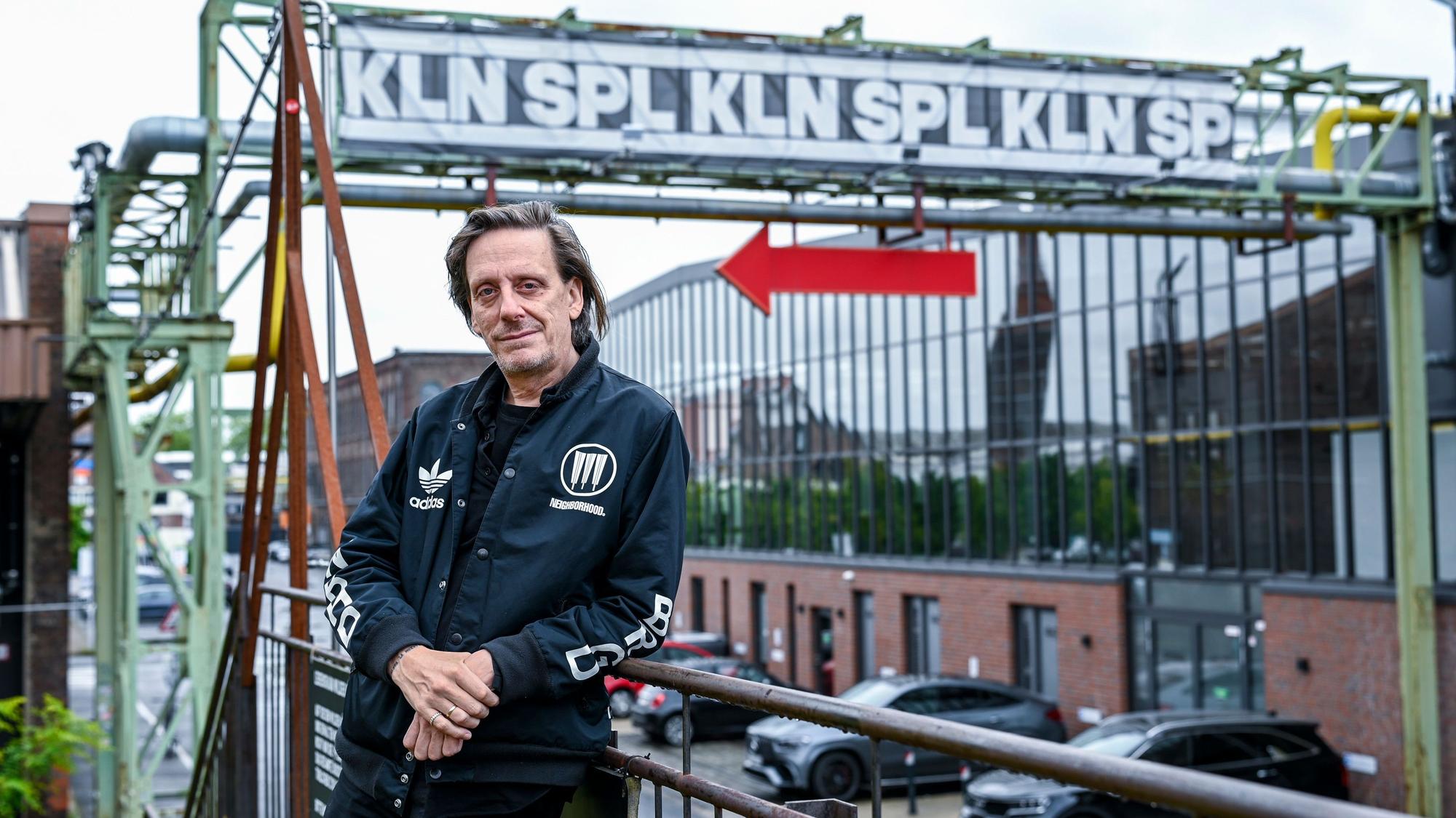 Ein Mann in schwarzer Jacke lehnt an einem Geländer. Über ihm ein Industriegerüst mit dem Schriftzug KLN SPL.