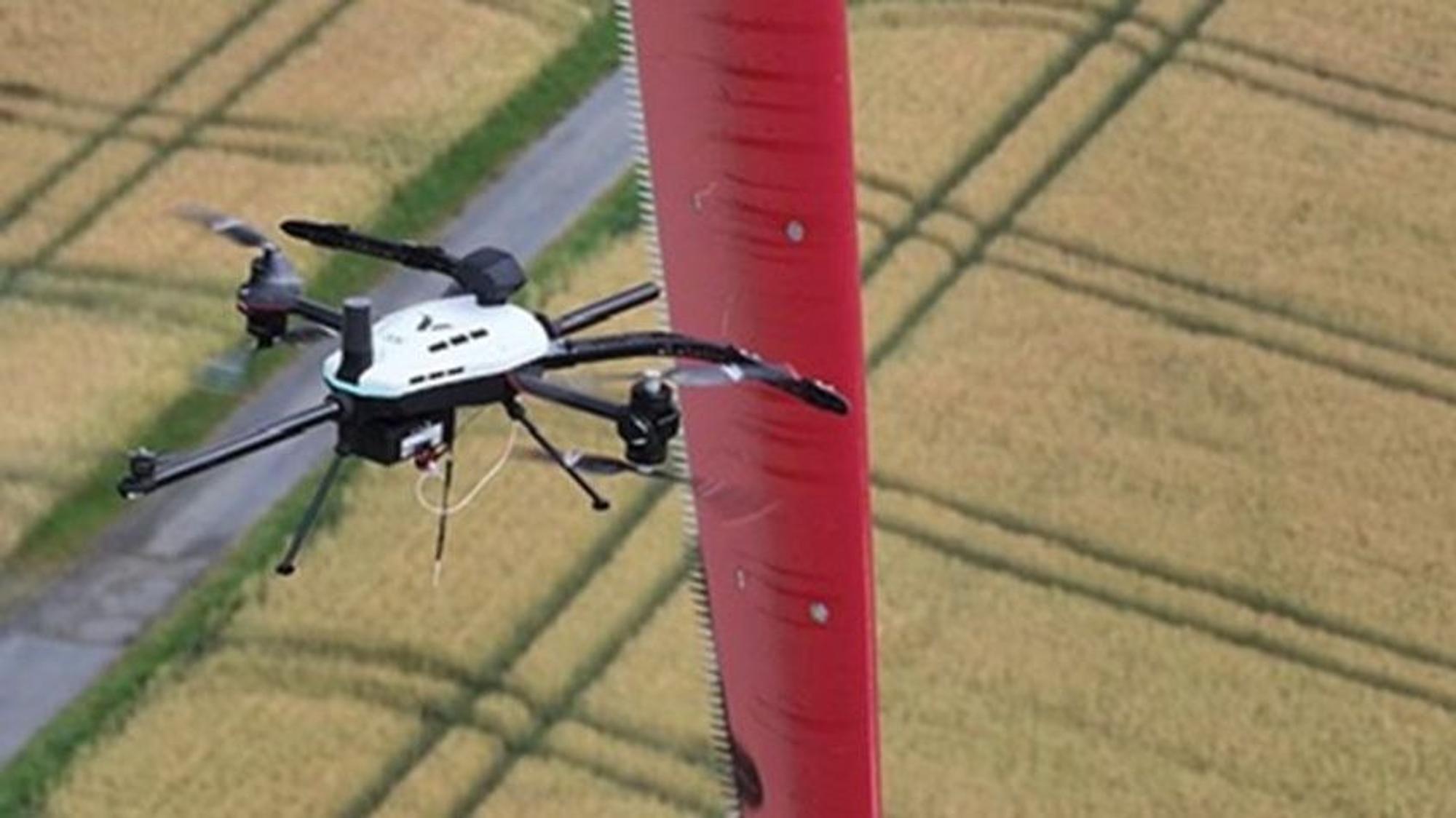 Dieses Foto zeigt, wie die schwarzweiße Drohne das rote Rotorblatt einer Windenergieanlage anfliegt.