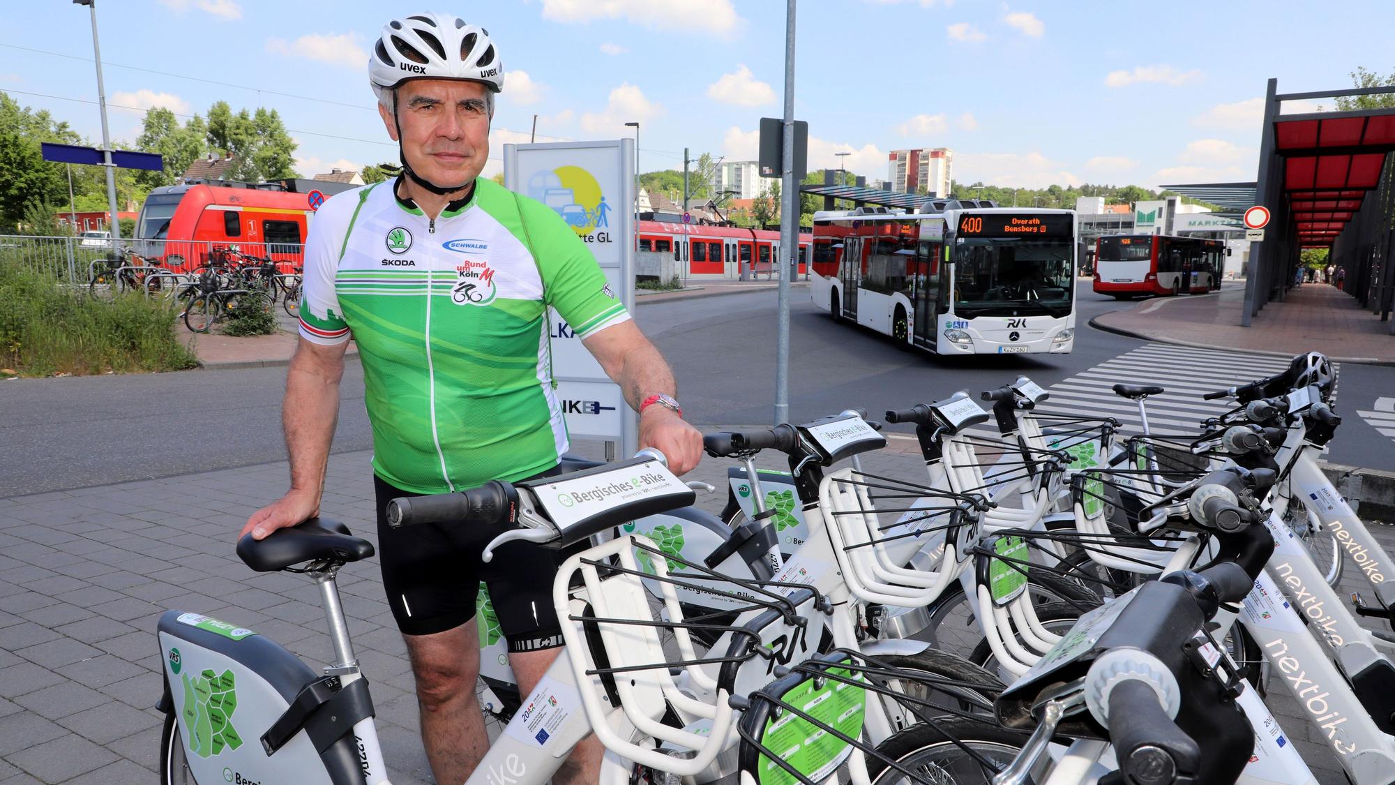 Der langjährige Streckenplaner von „Rund um Köln“ Werner Schneider steht an der Entleihstation des Bergischen E-Bikes am Bergisch Gladbacher S-Bahnhof. Im Hintergrund sind ein Bus und eine S-Bahn zu sehen.