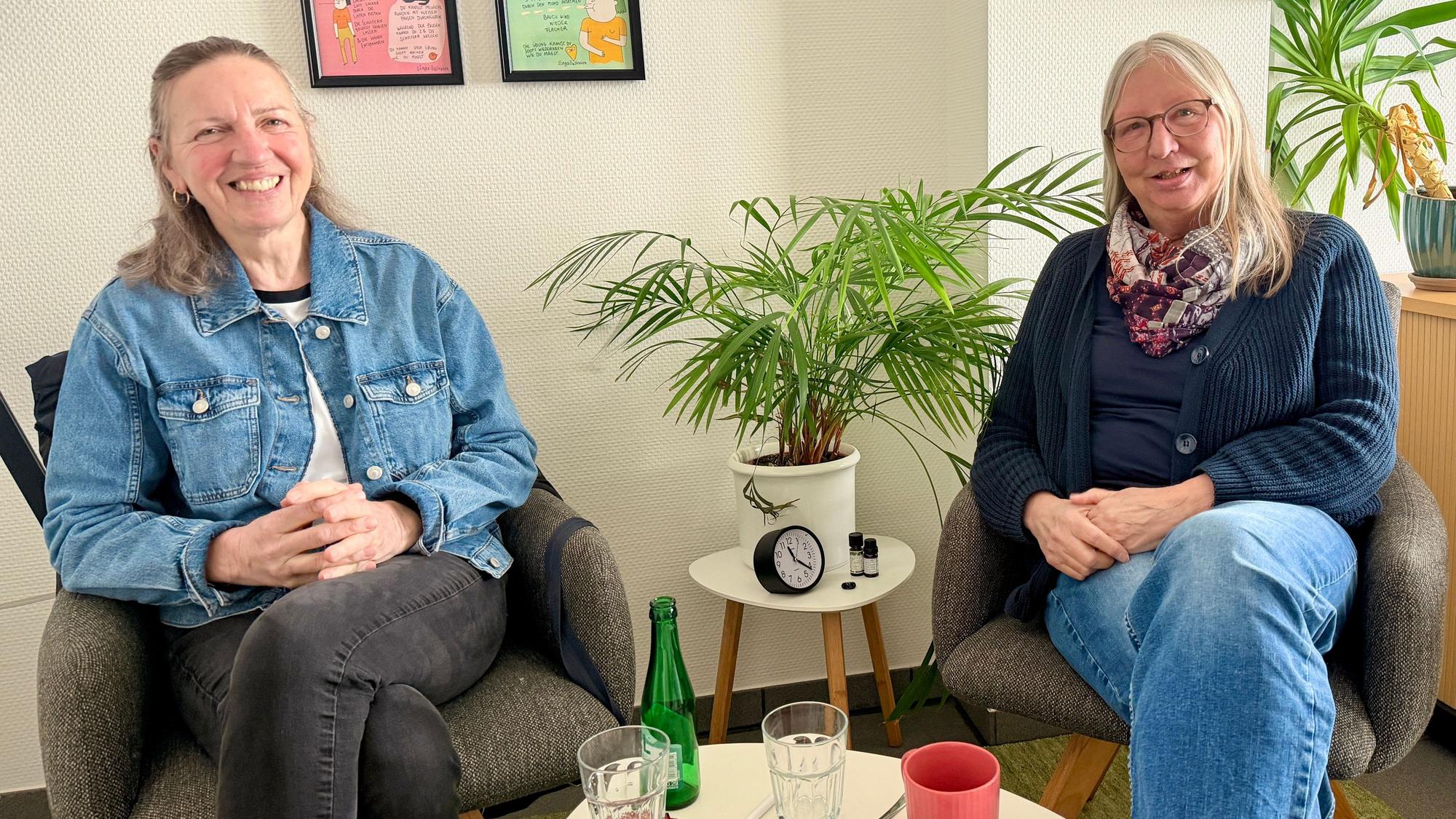 Die beiden Frauen sitzen auf Stühlen nebeneinander und lächeln in Richtung Kamera.