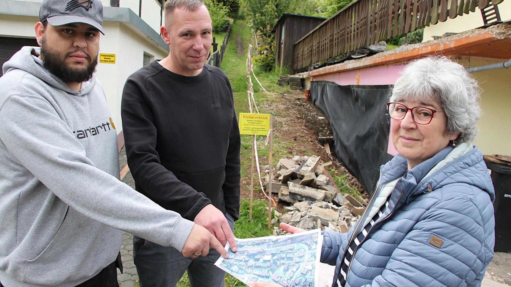 Zwei Männer und eine Frau stehen vor einem schmalen Wiesenweg zusammen. Sie schauen auf ein Luftbild, das die Frau in den Händen hält.