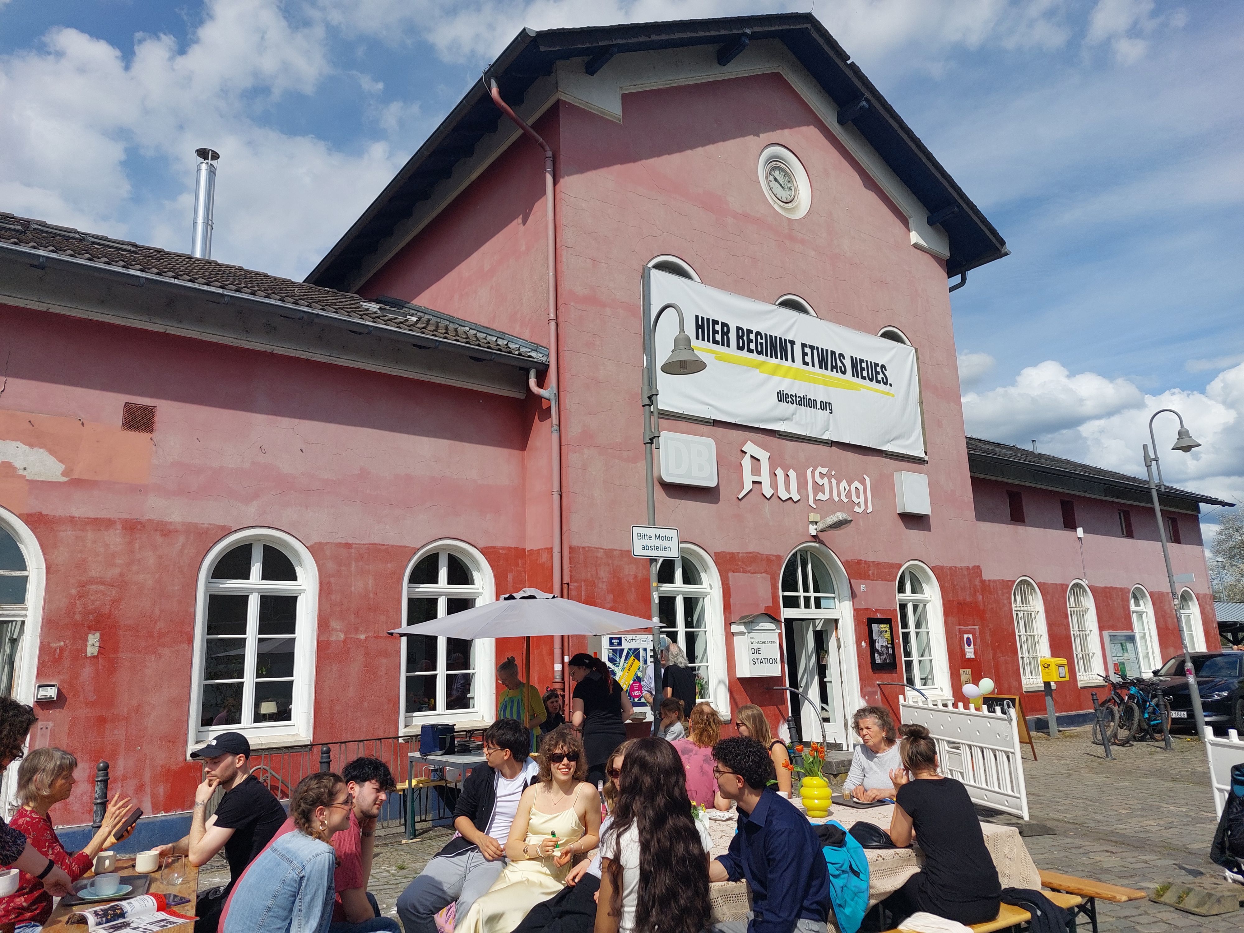 Ein alter Bahnhof, davor sitzen Menschen an Tischen