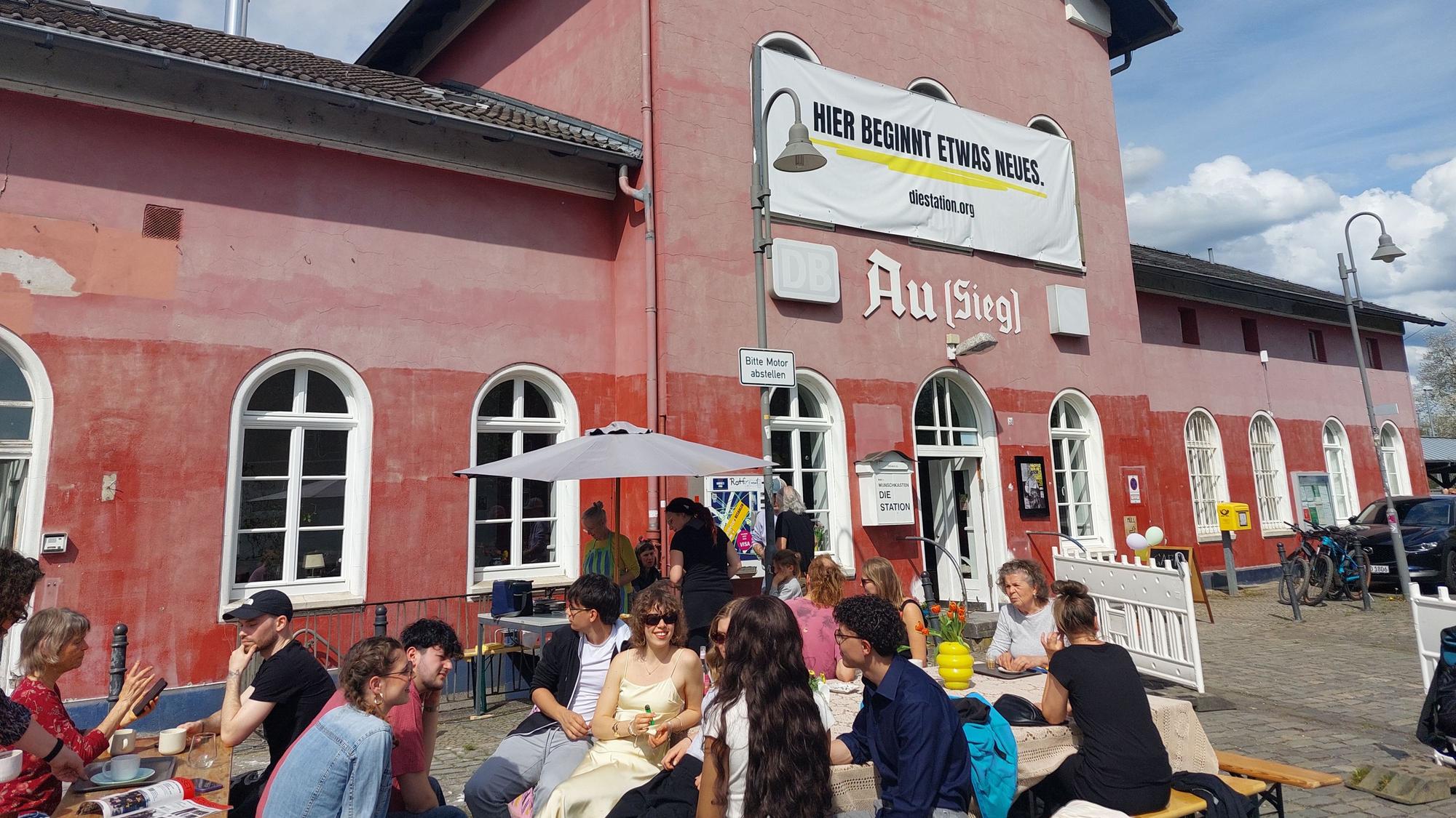 Ein alter Bahnhof, davor sitzen Menschen an Tischen
