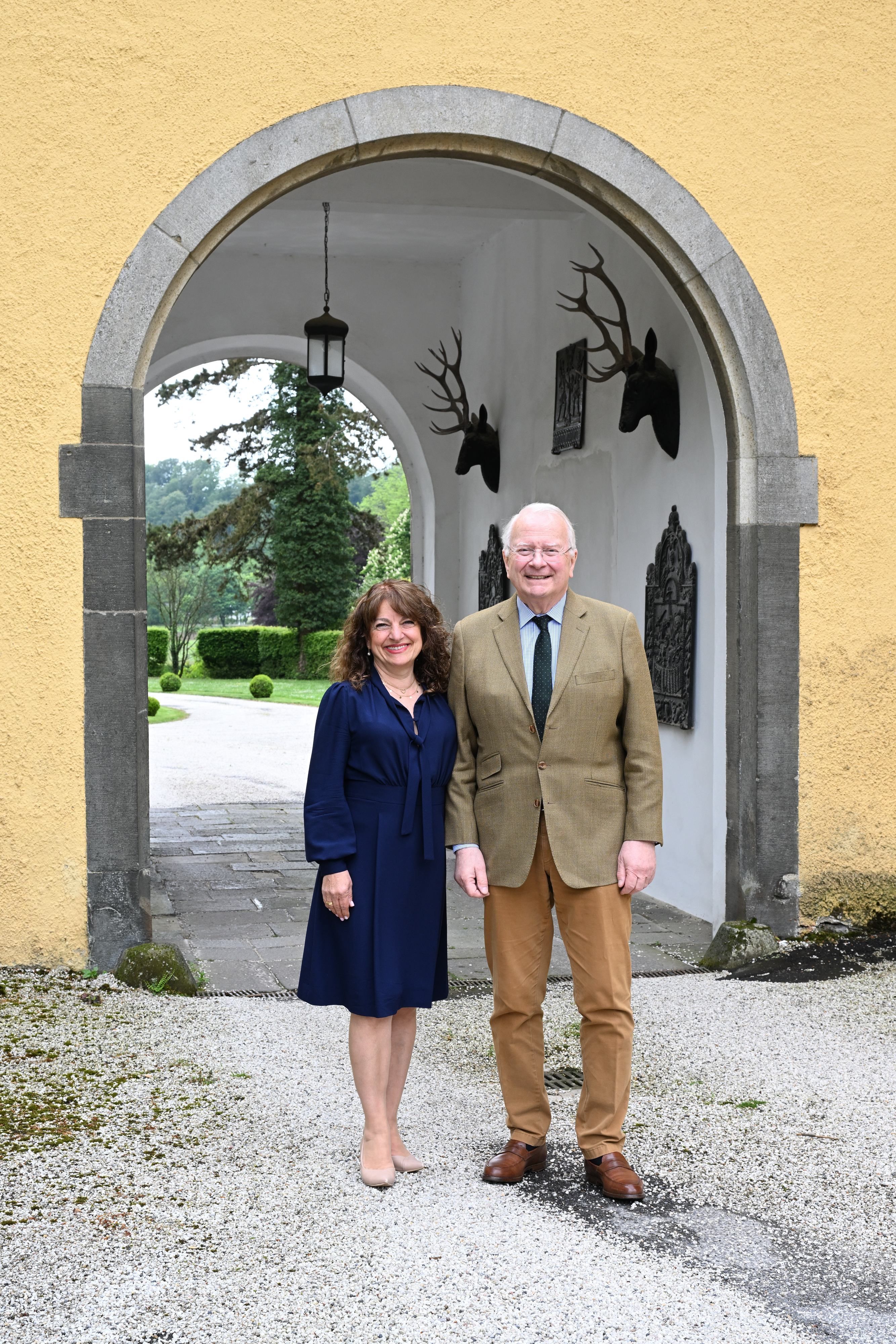 Eine Frau im dunkelblauen Kleid und ein Mann im Anzug stehen vor dem Torbogen eines Schlosses.