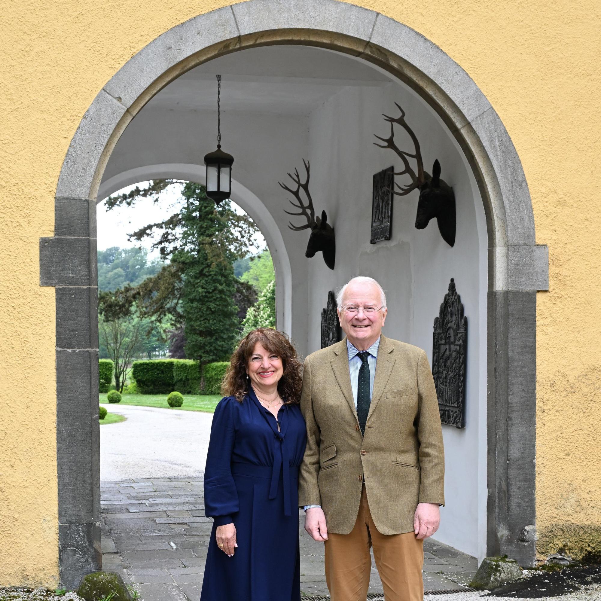 Eine Frau im dunkelblauen Kleid und ein Mann im Anzug stehen vor dem Torbogen eines Schlosses.
