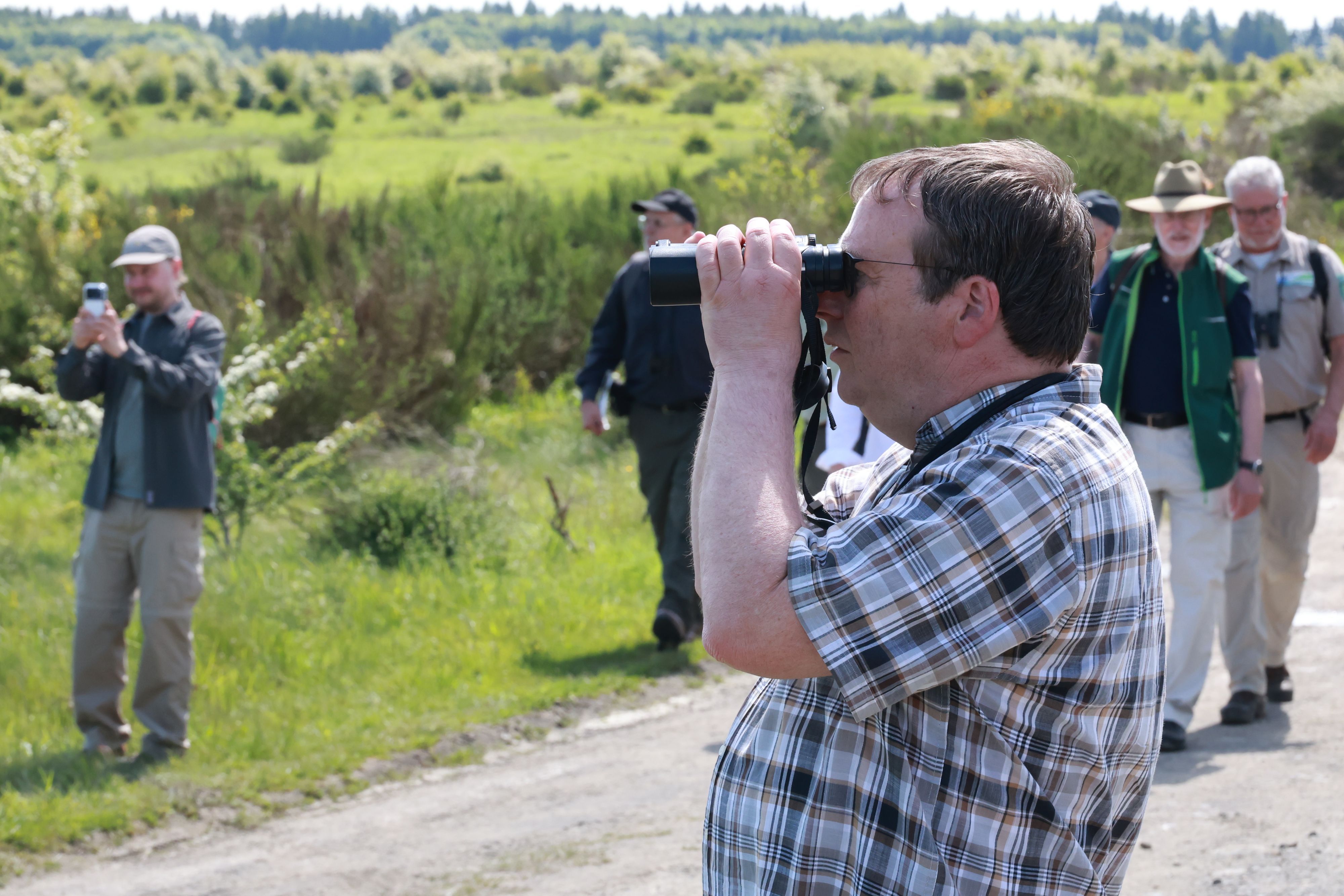Der Minister blickt durch ein Fernglas auf die Vegetationn neben dem Weg. Hinter ihm weitere Mitwanderer.