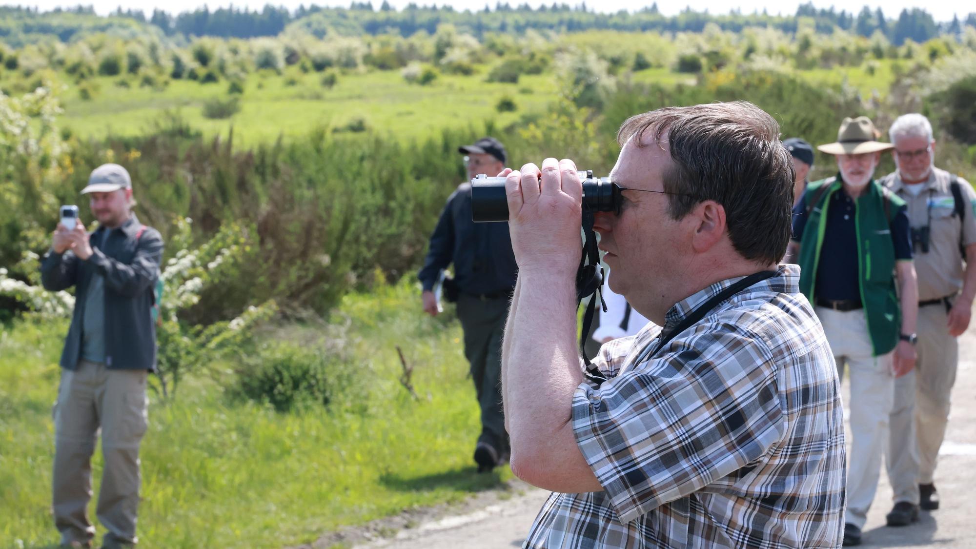 Der Minister blickt durch ein Fernglas auf die Vegetationn neben dem Weg. Hinter ihm weitere Mitwanderer.