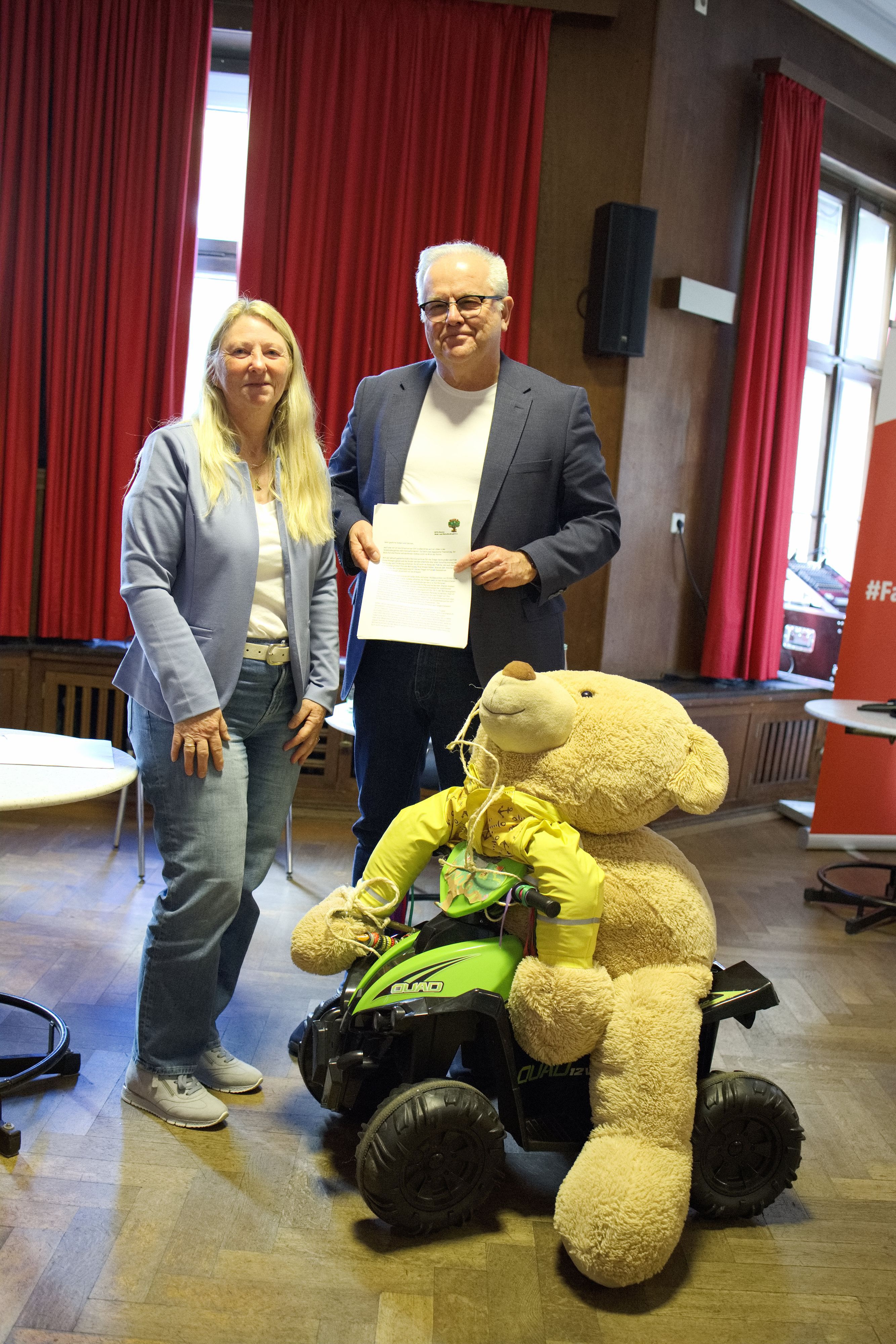 Auf einem großen Spielzeugtraktor sitzt ein großer Teddybär. Dahinter stehen ein Mann und eine Frau.