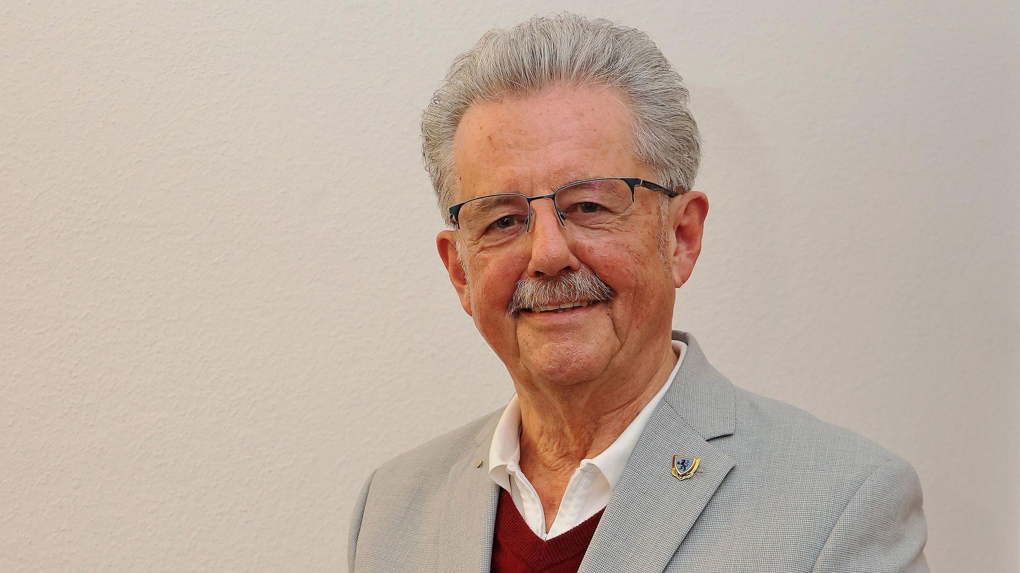 Ein Mann in grauem Sakko steht vor einer weißen Wand.