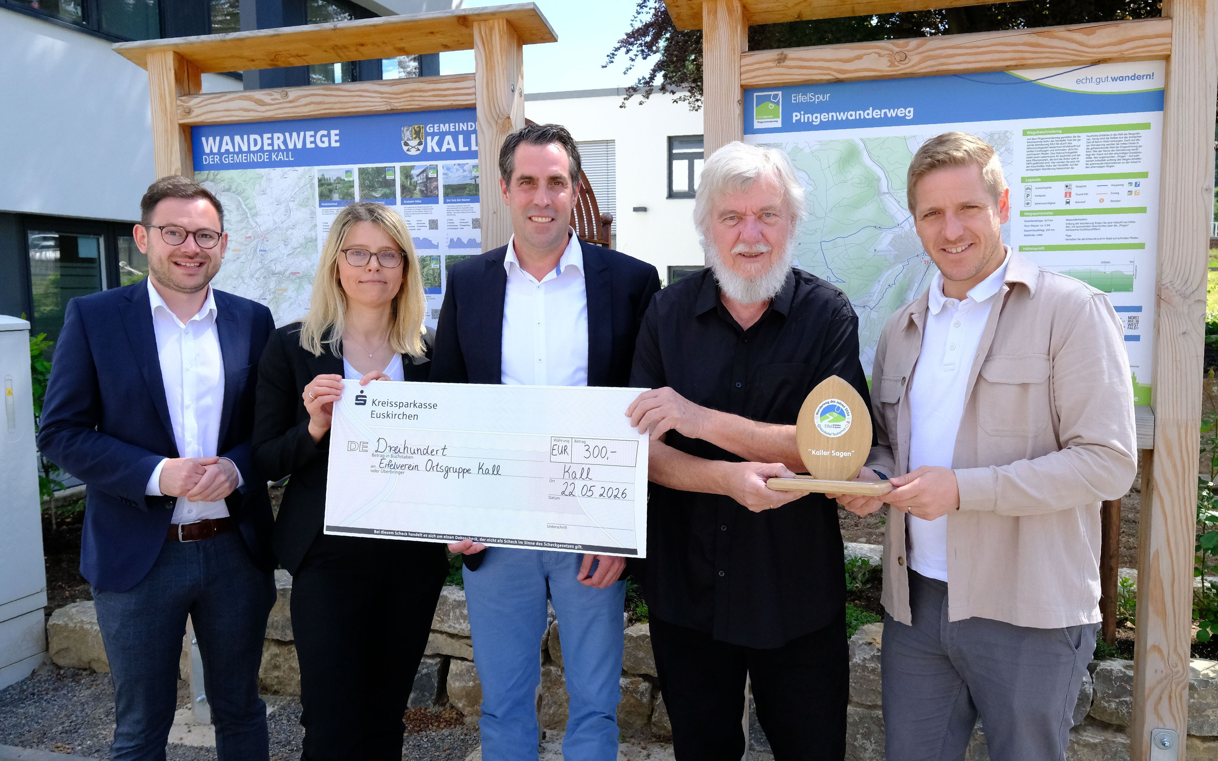 Die Genannten stehen mit einem überdimensionierten Scheck und einer Sieger-Trophäe vor zwei Wander-Infotafeln am Kaller Rathaus.