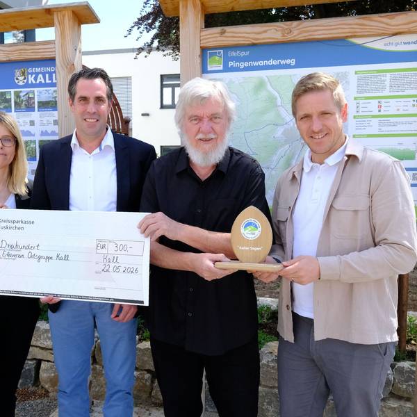Die Genannten stehen mit einem überdimensionierten Scheck und einer Sieger-Trophäe vor zwei Wander-Infotafeln am Kaller Rathaus.
