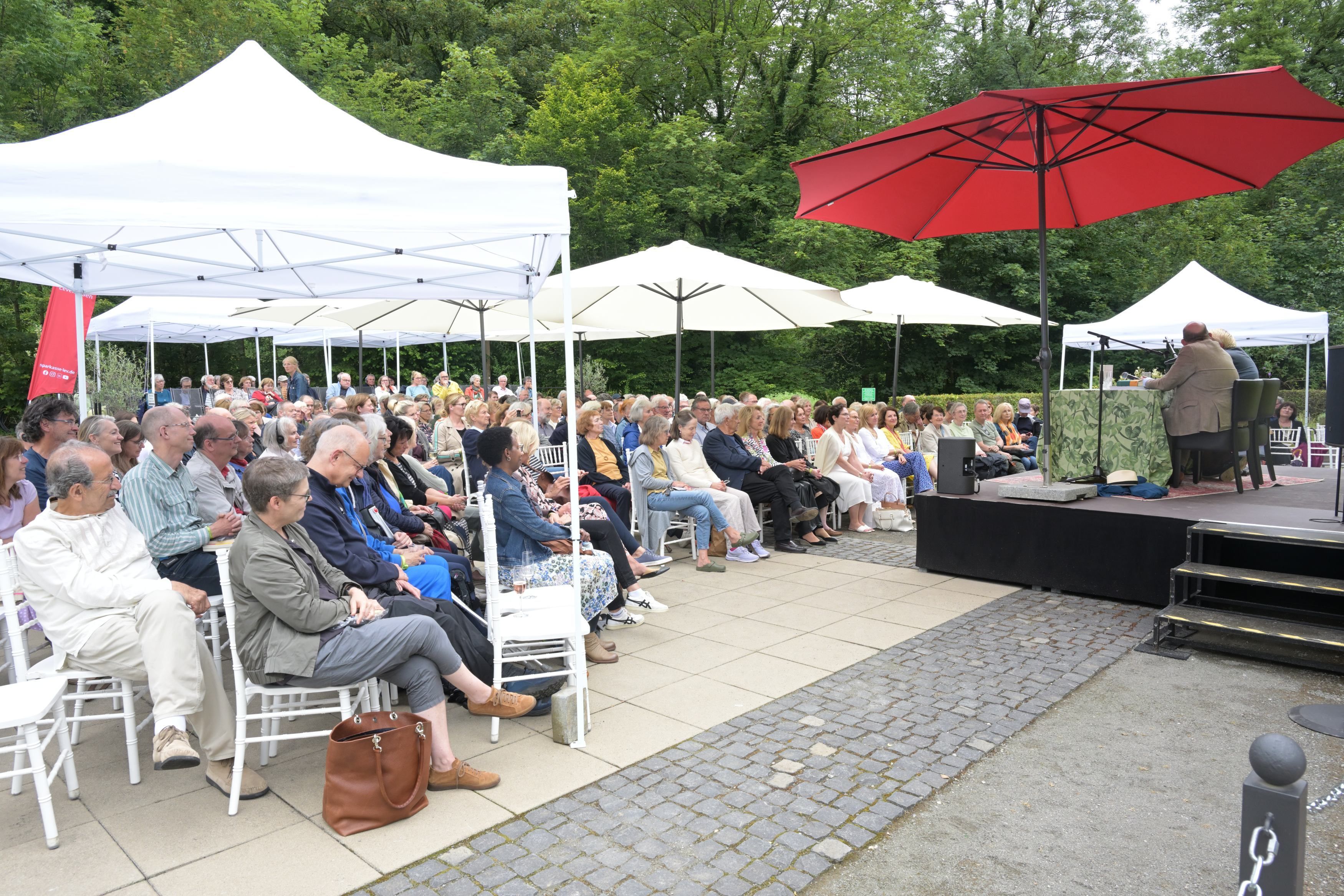 Eine Open-Air-Bühne mit weißen und roten Sonnenschirmen und Publikum auf weißen Stühlen.