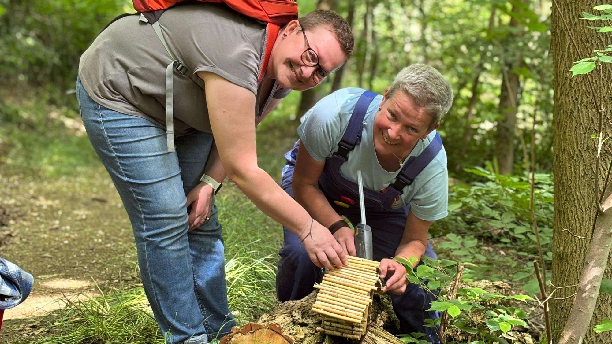 Die Obengenannten kleben ein Häuschen in einem Wald mit einer Klebepistole fest und lächeln in die Kamera.