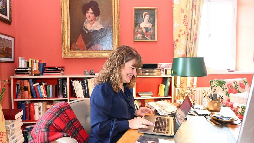 Eine Frau sitzt an einem Laptop in einem Arbeitszimmer mit historischen Ölporträts an den terrakottafarbenen Wänden.
