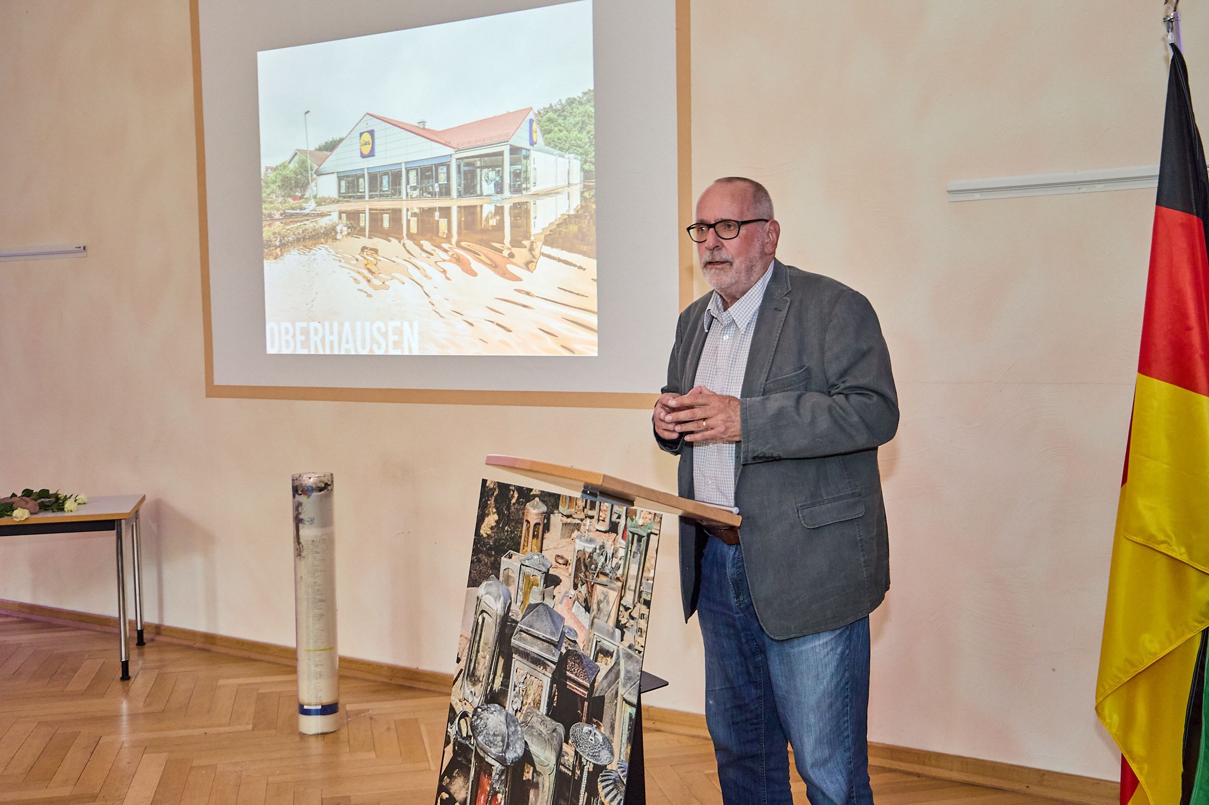 Klaus Jansen steht an einem Rednerpult, links von ihm steht die Flutkerze und an der Wand dahinter wird eine Präsentation gezeigt.