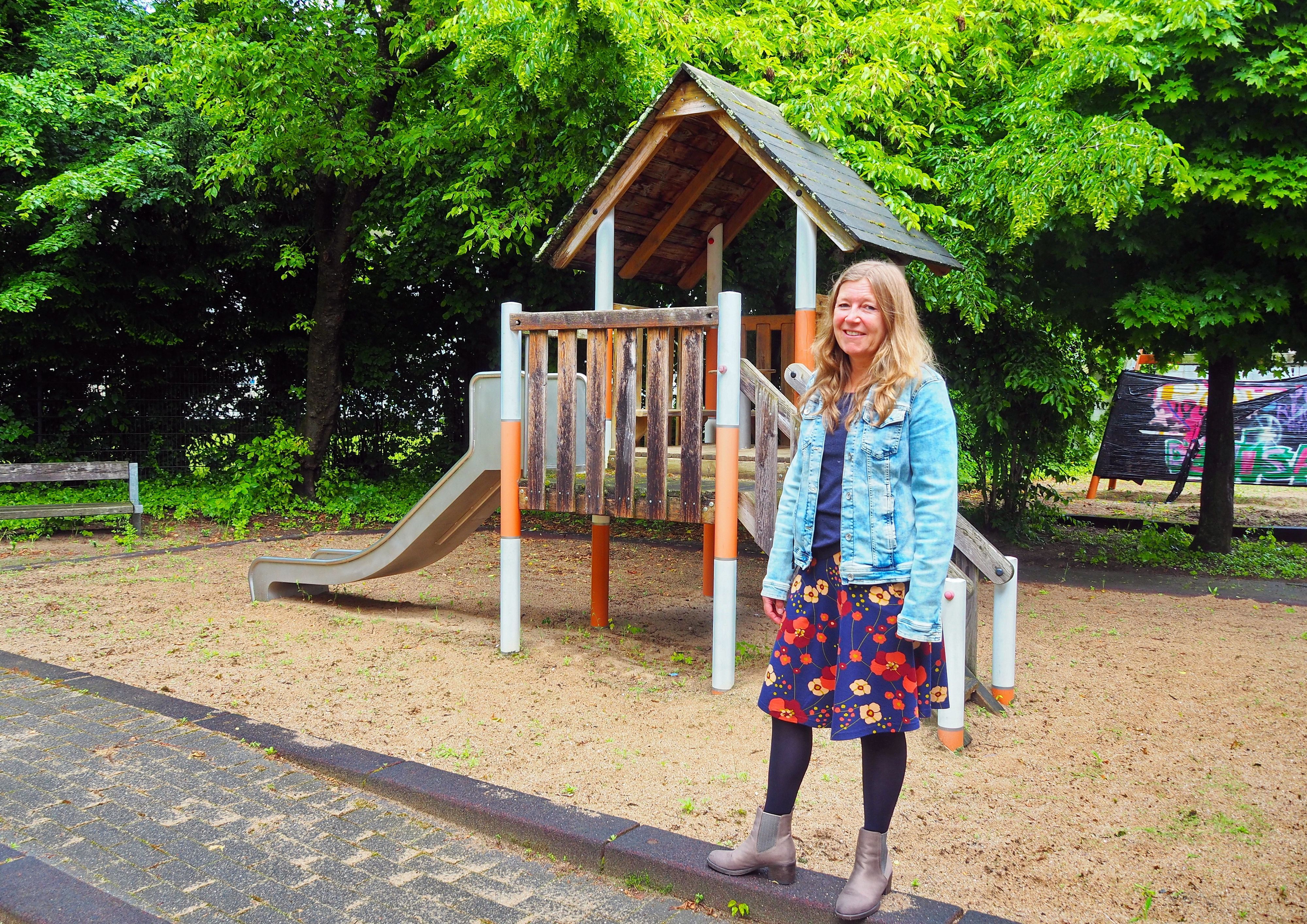 Eine Frau in Jeansjacke und Blumenkleid steht vor einem Spielehaus auf einer Sandfläche eines Spielplatzes.