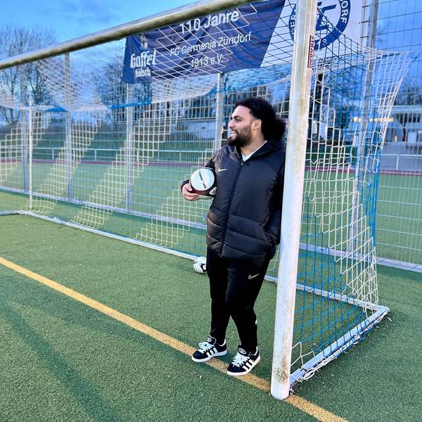Ein Mann mit dunklem Haar und Trainingsklamotten steht mit Fußball unter dem Arm an einen Pfosten eines Fußballtors gelehnt.