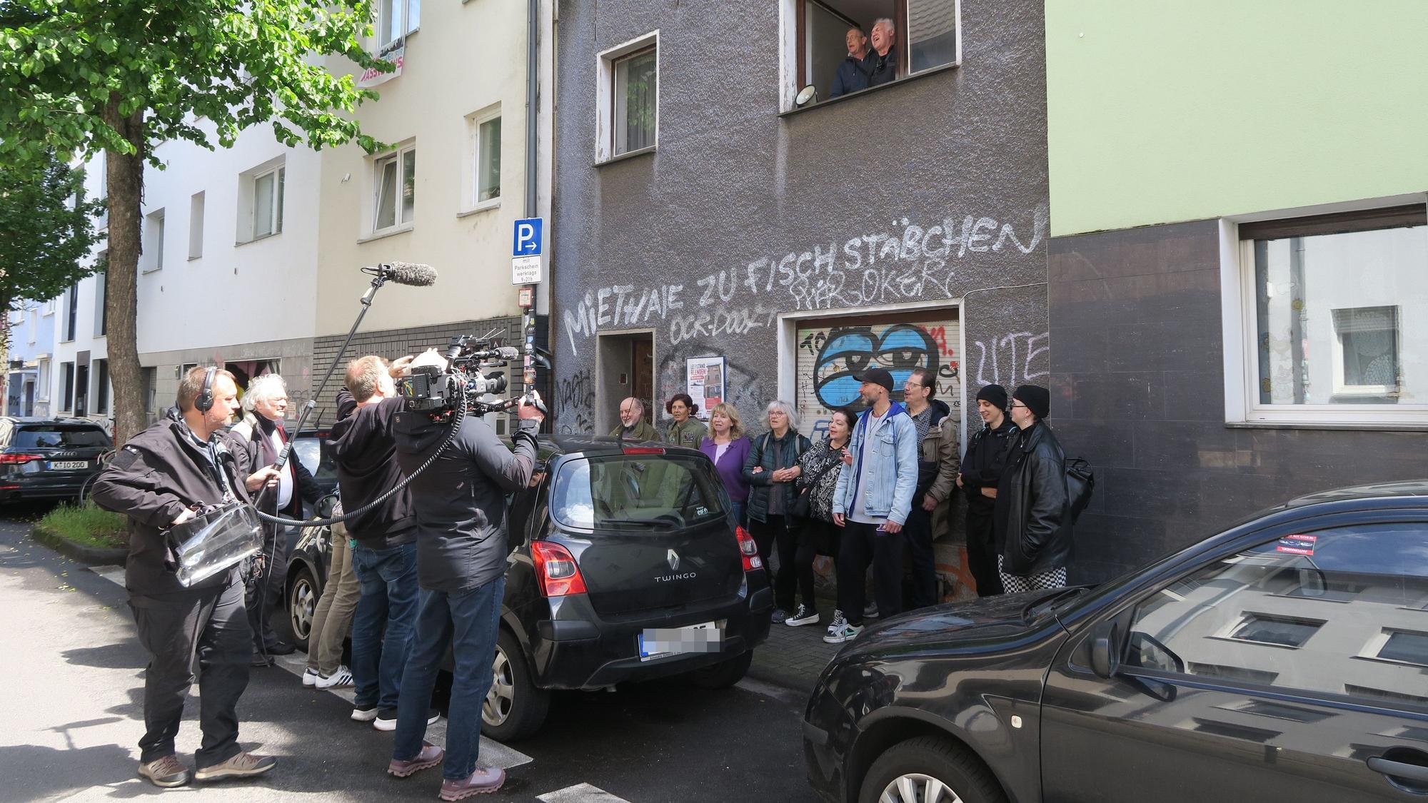 Ein Kamerateam filmt eine Menschengruppe, die sich vor einem Haus versammelt hat.
