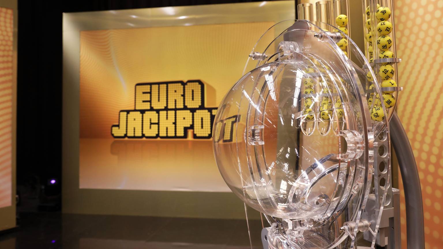 Das Bild zeigt die Ziehungsmaschine für die Eurojackpot-Lotterie. Im Hintergrund ist groß das goldene „EURO JACKPOT“-Logo auf einer beleuchteten Wand zu sehen.