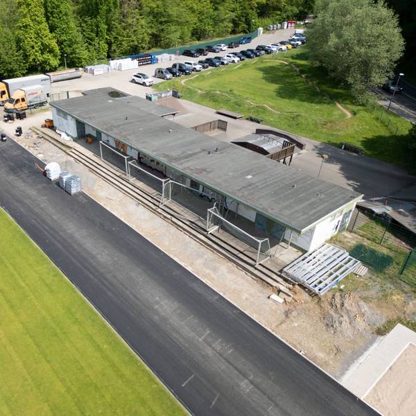 Ein Sportplatz im Umbau. Grüner Rasen ist schon verlegt, die Tartanbahn wird gerade gebaut. Dahinter ein verhülltes Gebäude. Ein Flachbau ist marode.