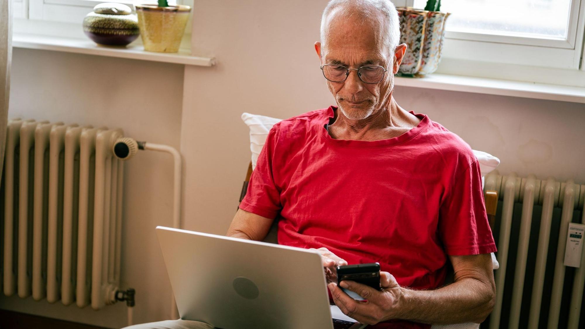 Ein Senior sitzt mit Laptop und Smartphone auf einem Sessel