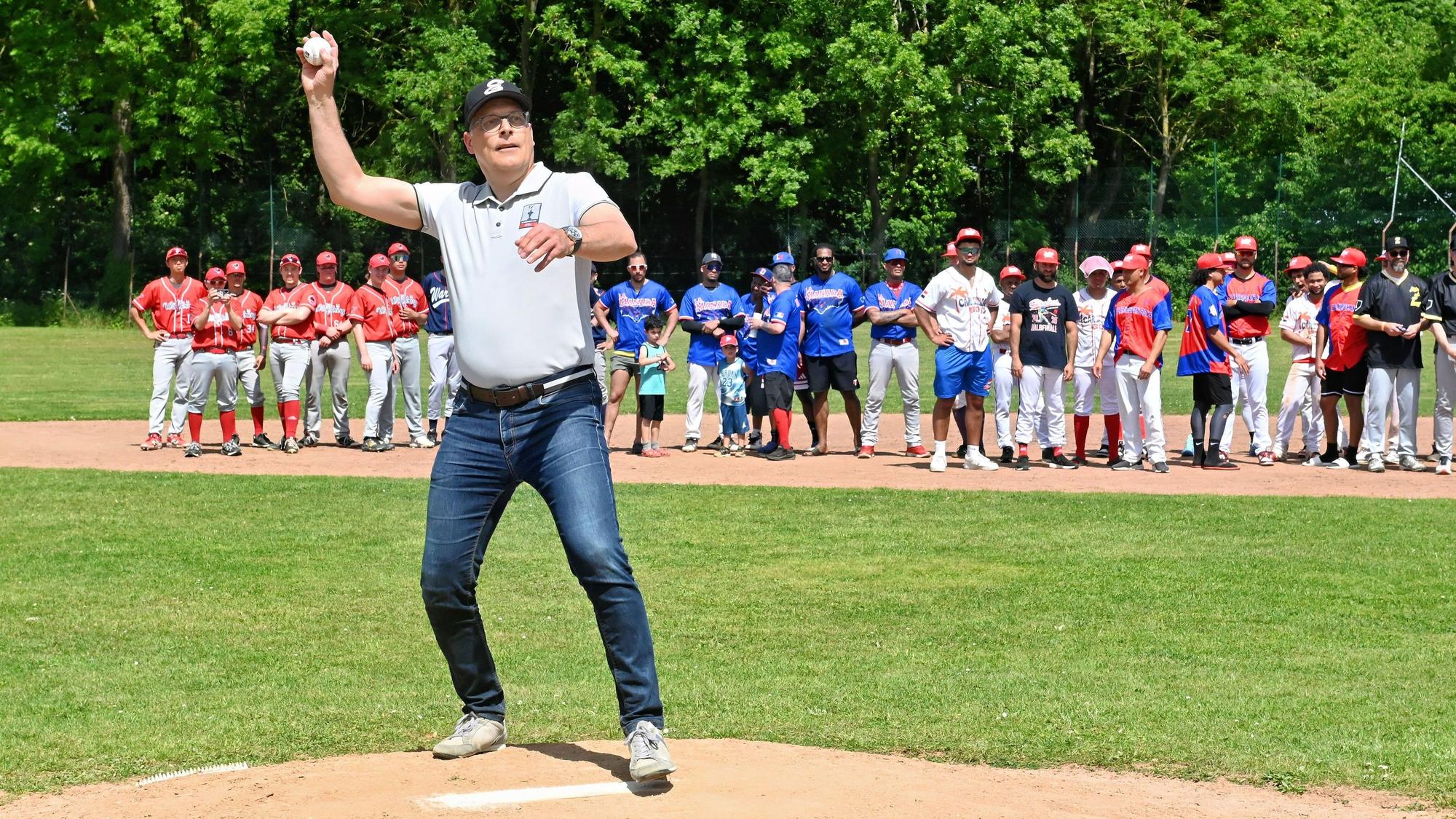 Bürgermeister Ulf Hürtgen steht auf dem Pitcherhügel und wirft einen Ball, im Hintergrund schauen mehrere Mannschaften zu.
