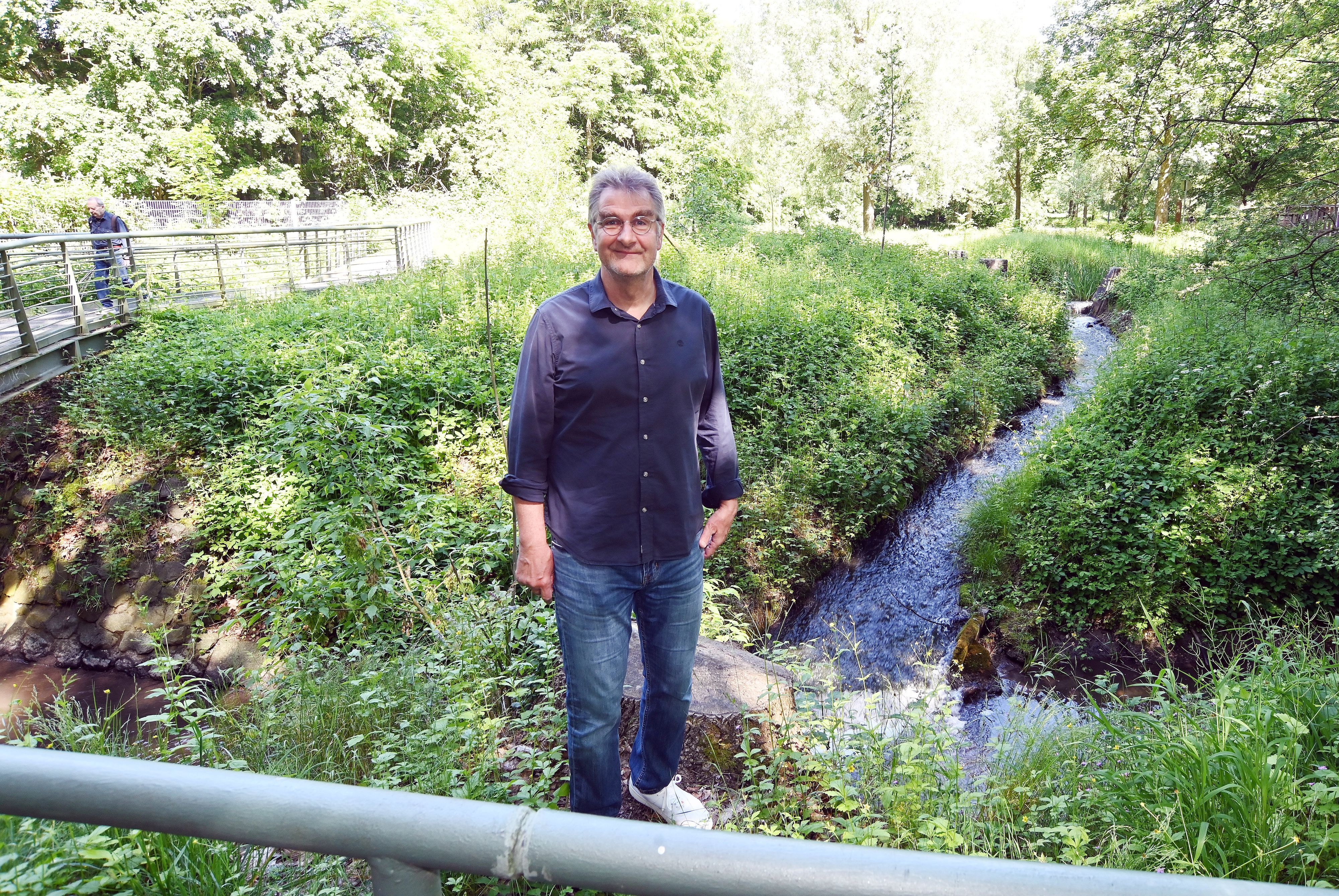 Ein Mann im blauen Hemd und Jeans steht neben einem Bachlauf.