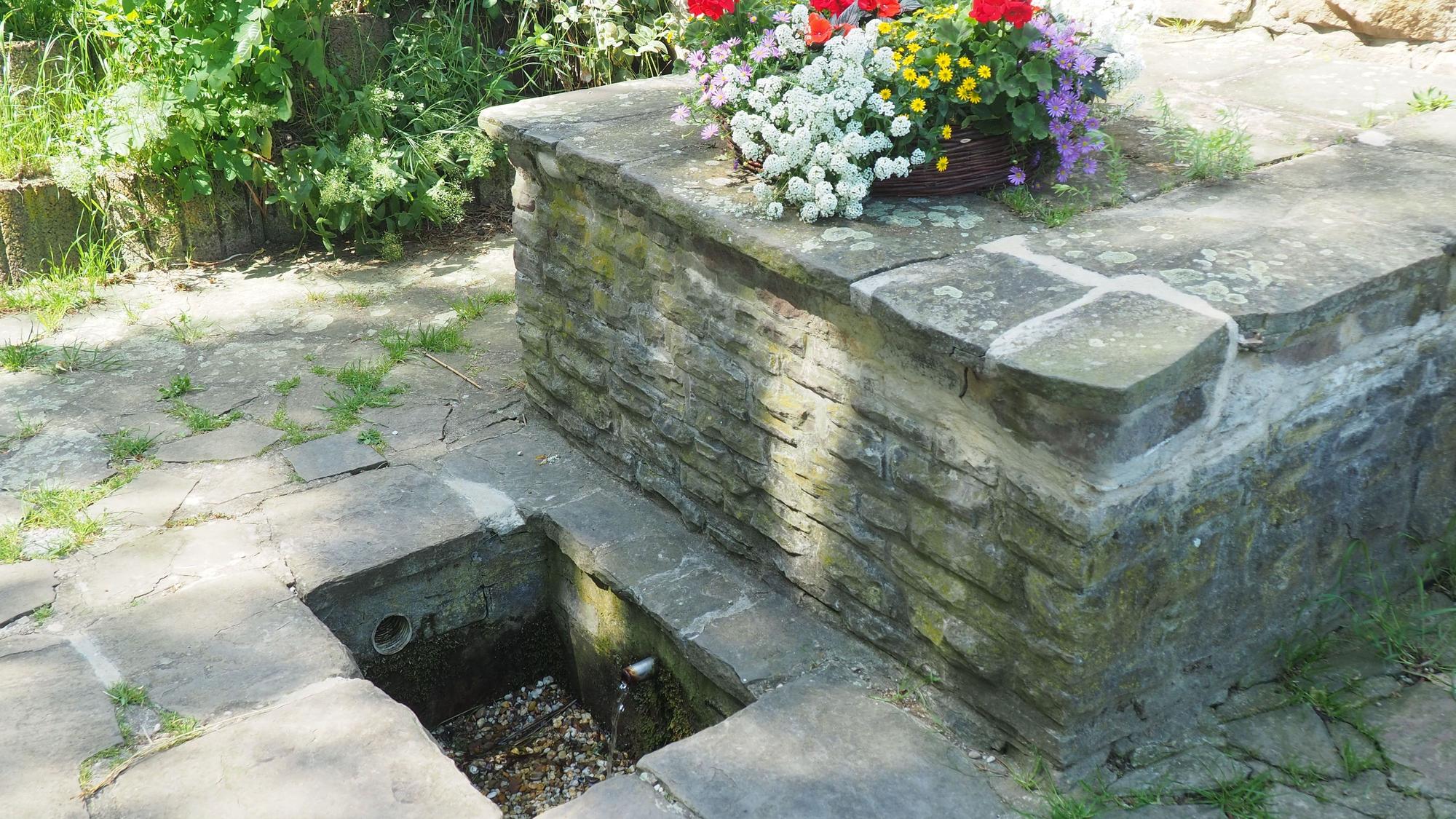 In einem kleinen, viereckigen Becken im Boden rinnt Wasser aus einem Rohr. Dahinter steht eine mit Blumen bepflanzte Schale.
