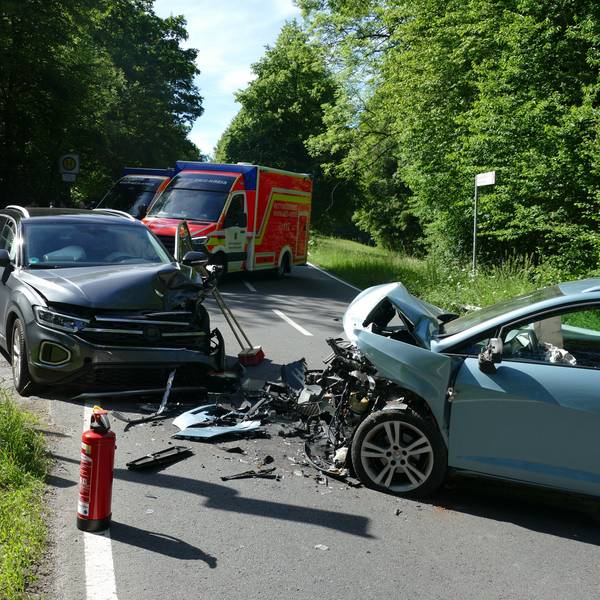 Zwei frontal zusammengestoßene Autos, zwischen ihnen Fahrzeugteile. Im Hintergrund steht ein Rettungswagen.