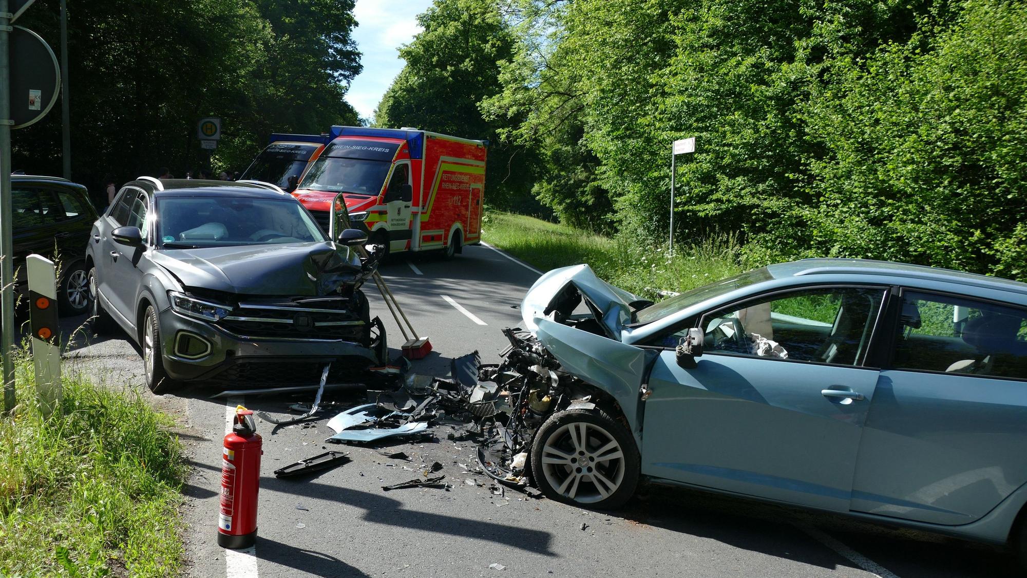 Zwei frontal zusammengestoßene Autos, zwischen ihnen Fahrzeugteile. Im Hintergrund steht ein Rettungswagen.