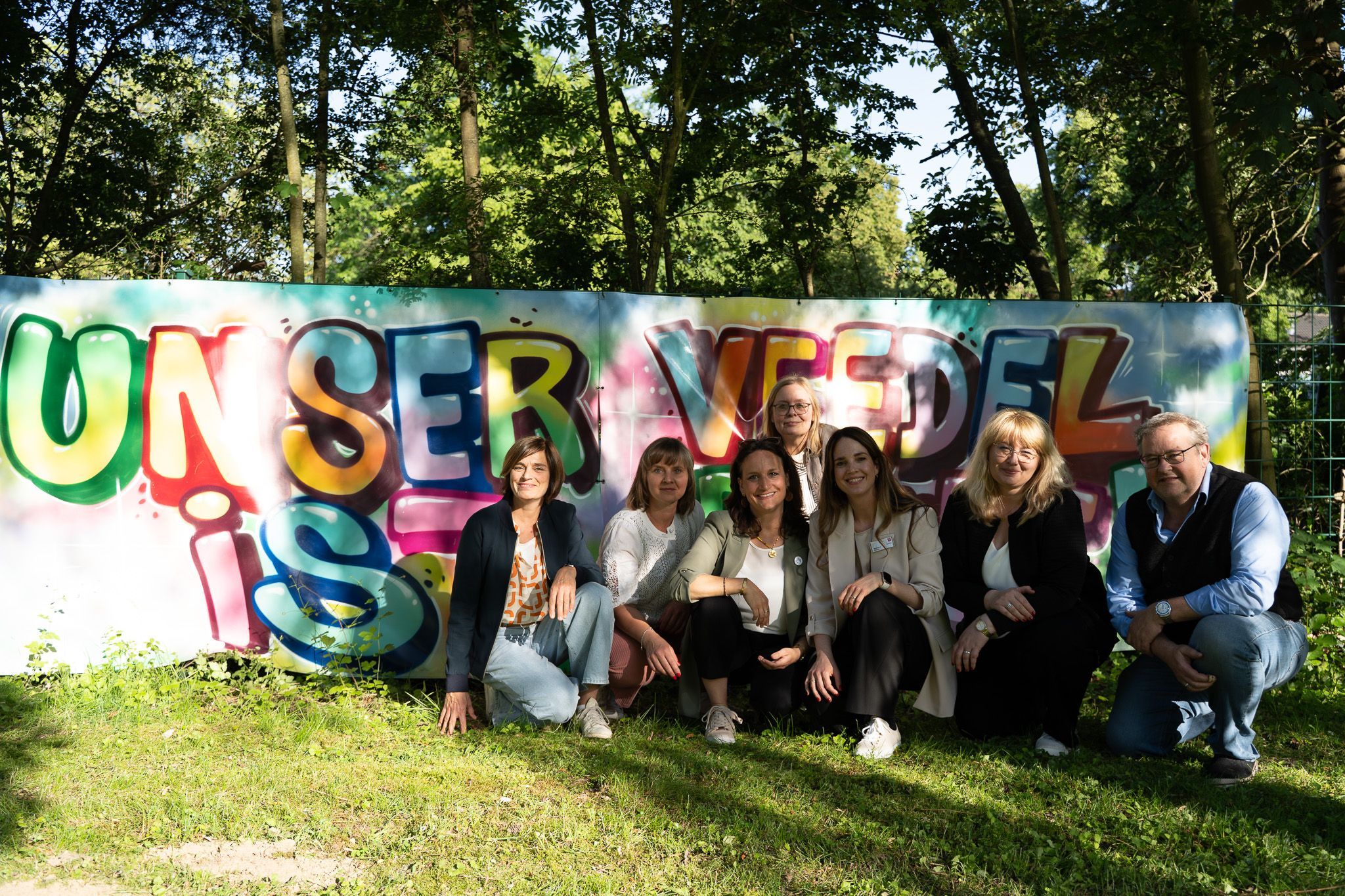 Akteure der Quartiersentwicklung im Kölner Süden hocken vor einer Wand, auf der ein Graffiti gesprüht ist: Unser Veedel ist bunt.