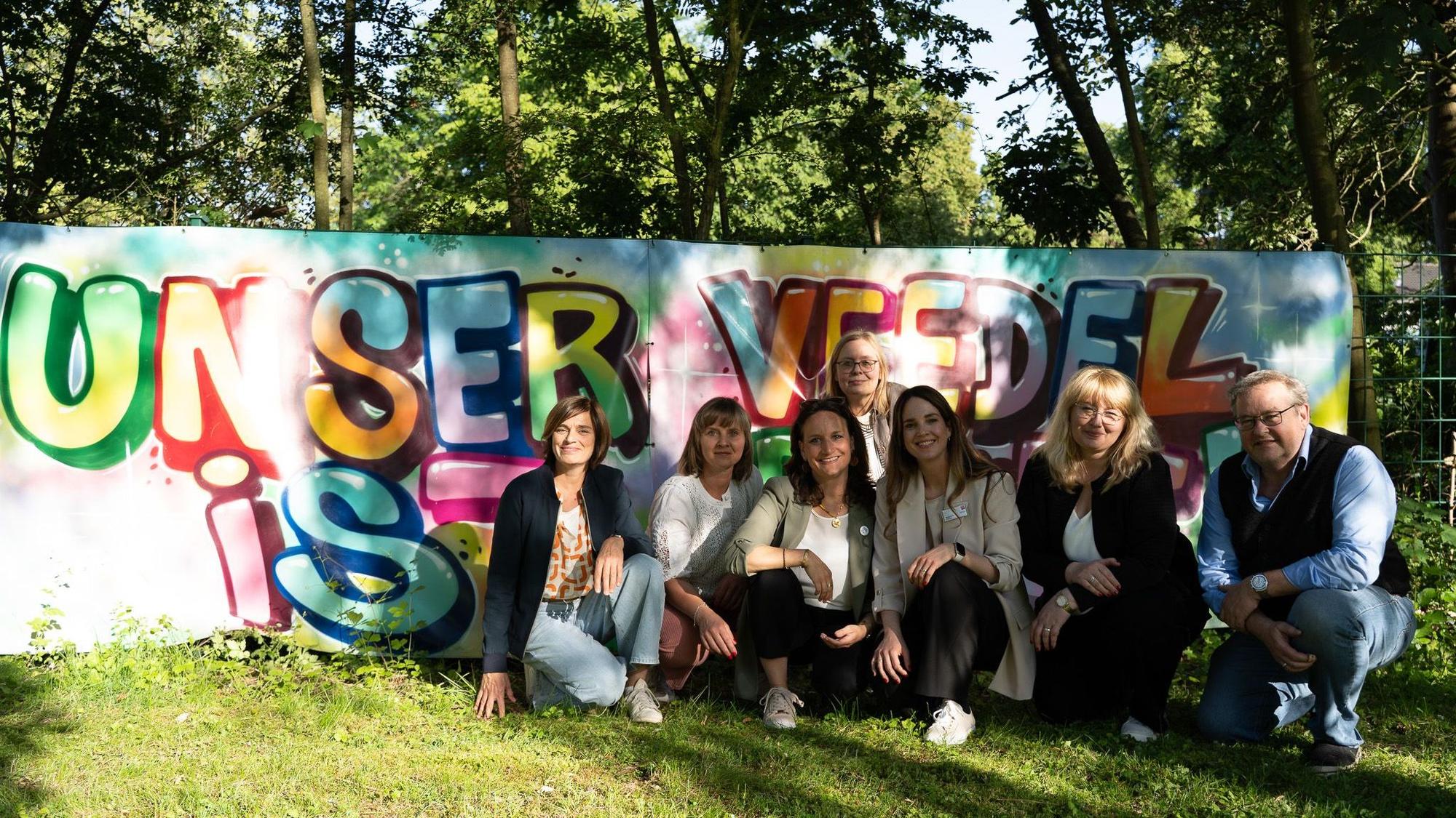 Akteure der Quartiersentwicklung im Kölner Süden hocken vor einer Wand, auf der ein Graffiti gesprüht ist: Unser Veedel ist bunt.