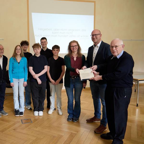 Mehrere Jugendliche und Erwachsene stehen nebeneinander. Ein Mann übergibt einem anderen und einer Frau ein Schriftstück.