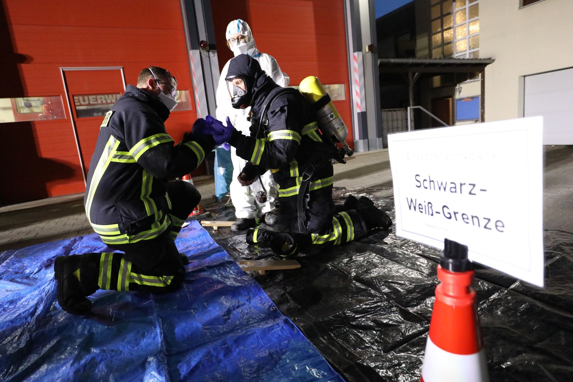 Ein knieender Feuerwehrmann legt mit Unterstützug seine Einsatzhkleidung ab, rechts ein Schild „ Schwarz-Weiß-Grenze“.