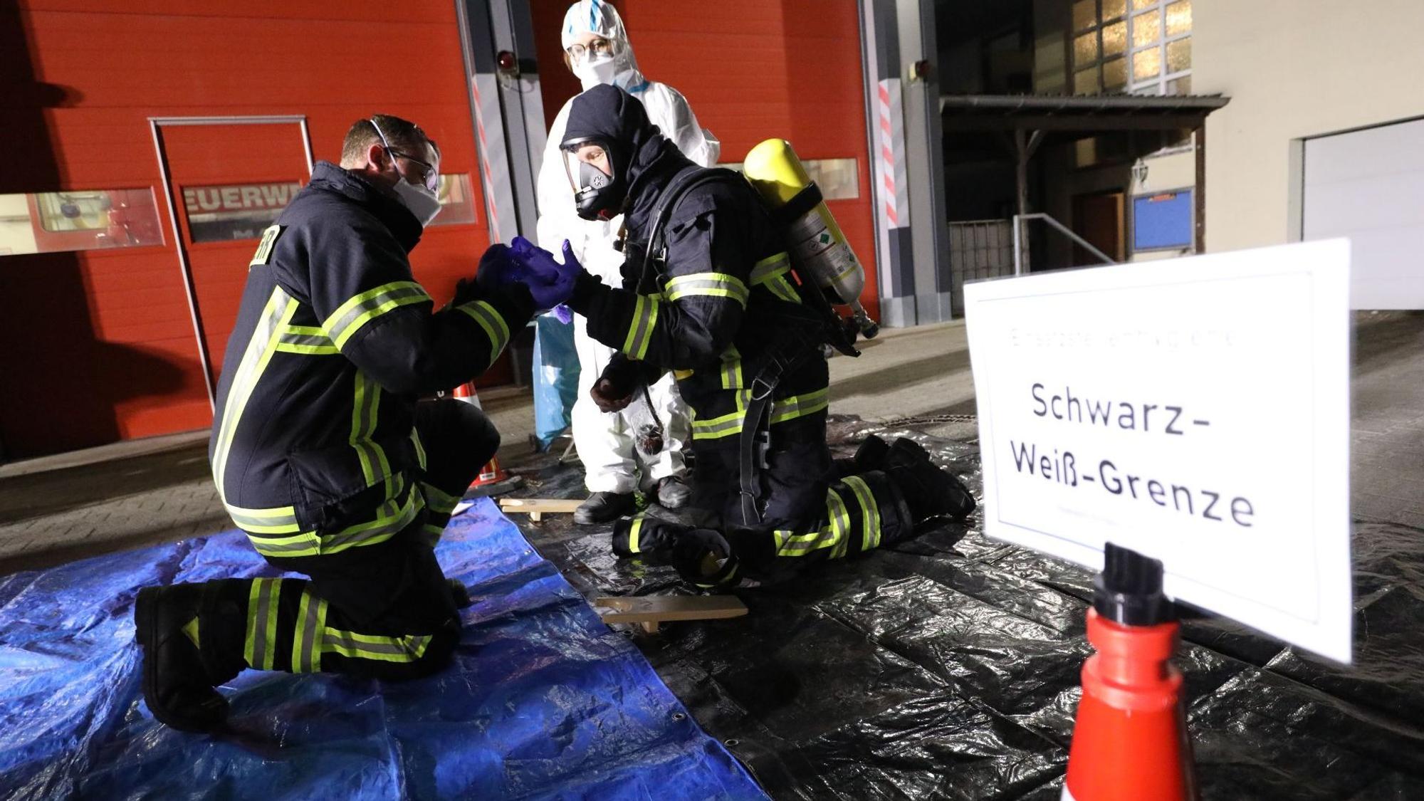 Ein knieender Feuerwehrmann legt mit Unterstützug seine Einsatzhkleidung ab, rechts ein Schild „ Schwarz-Weiß-Grenze“.