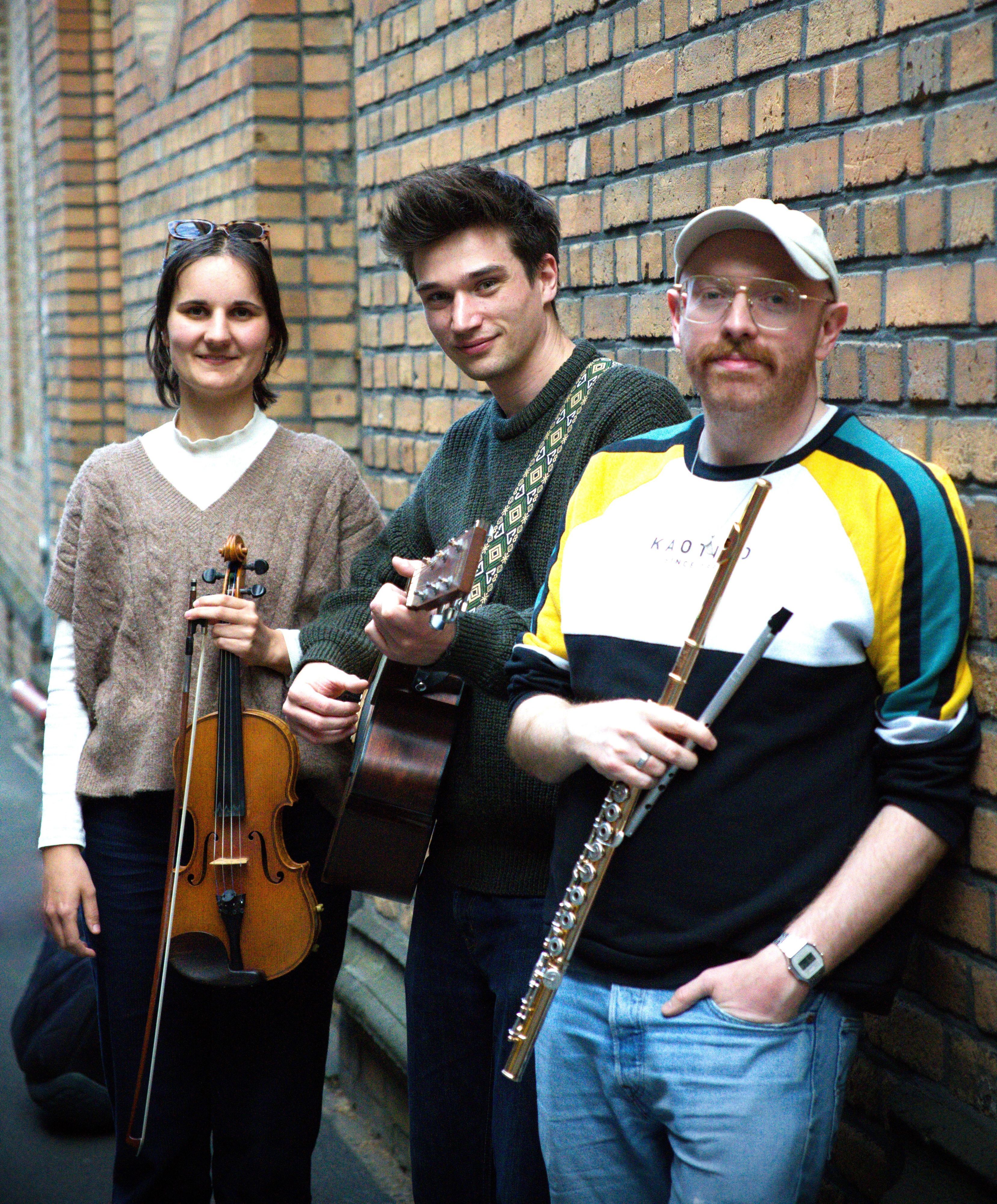 Die Obengenannten stehen mit eine Geige, Gitarre und Flöte an einer Backsteinwand.