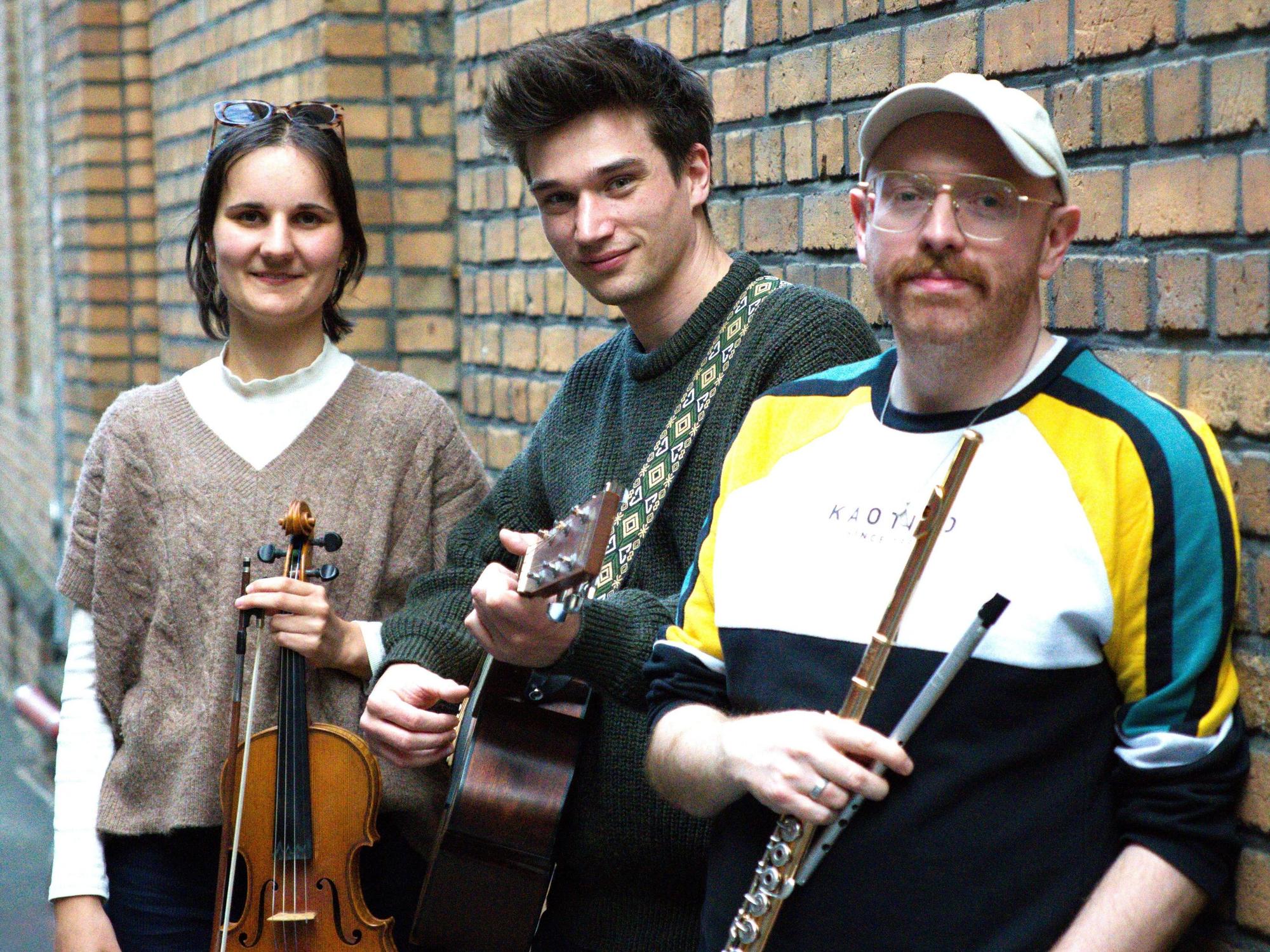 Die Obengenannten stehen mit eine Geige, Gitarre und Flöte an einer Backsteinwand.