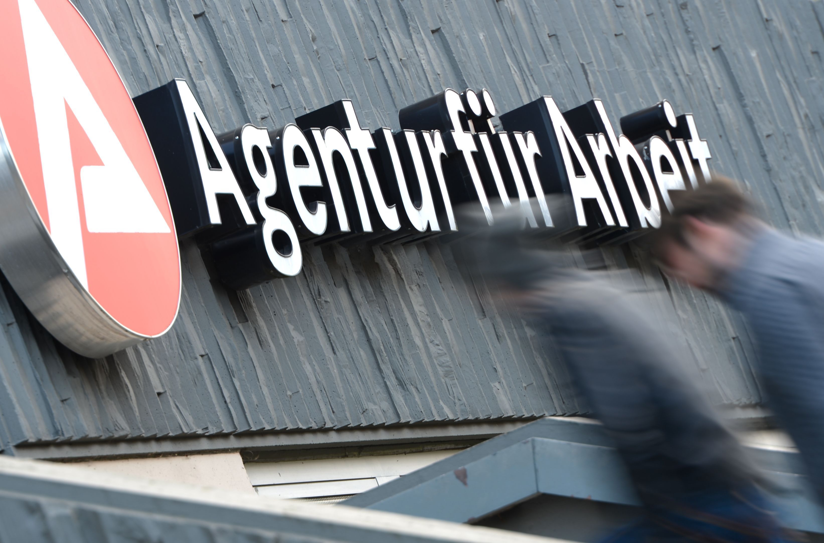 Das Symbolfoto zeigt zwei Männer vor einem Schild einer Agentur für Arbeit.
