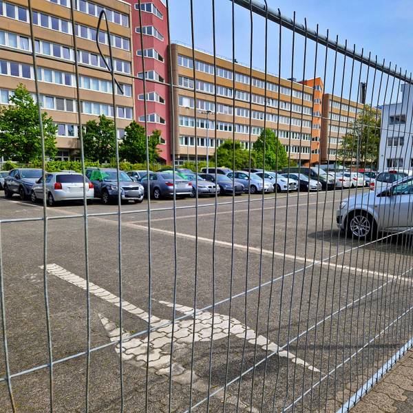 Hinter einem Bauzaun ist ein Parkplatz mit vielen abgestellten Autos.