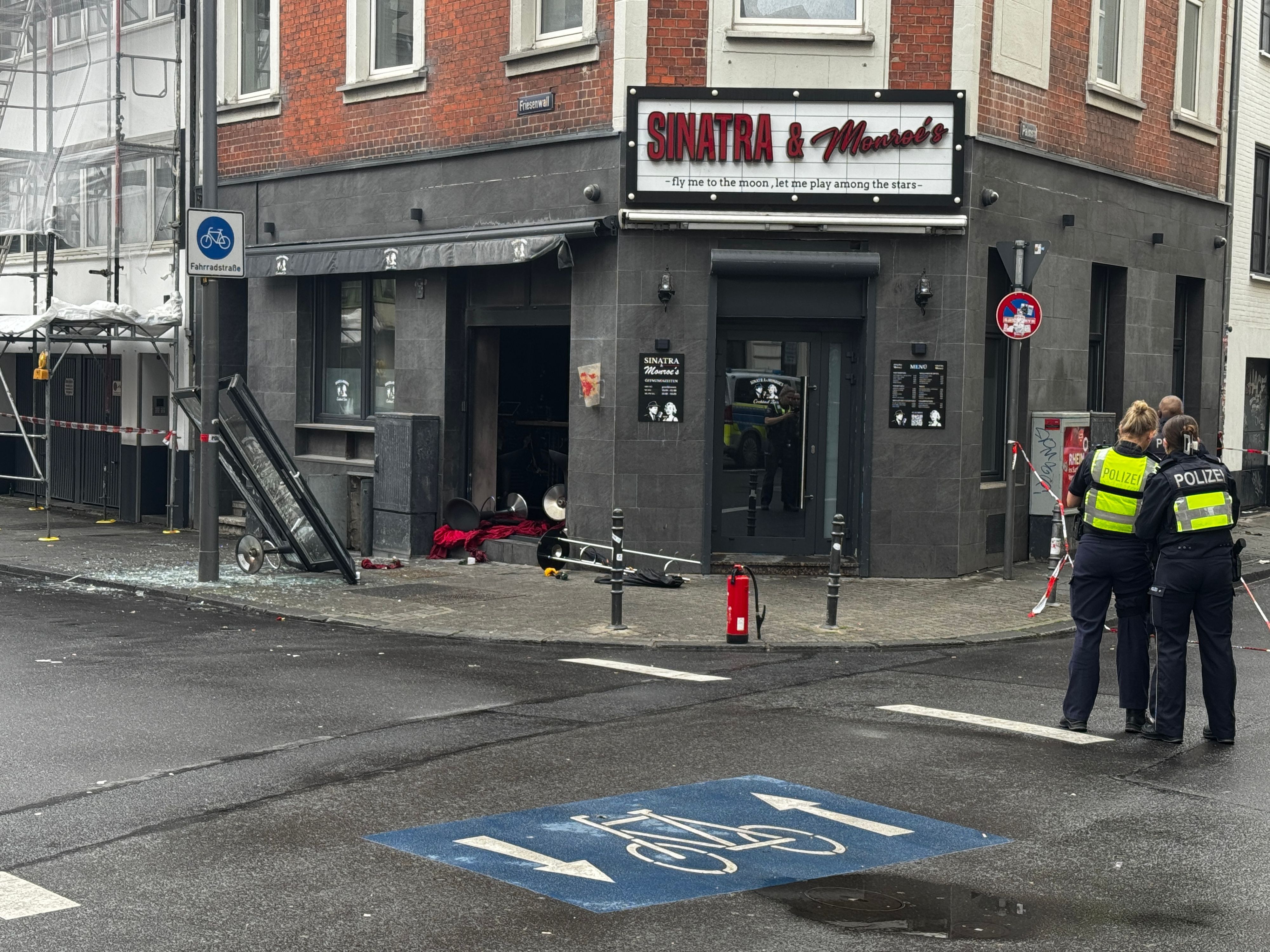 Blick auf die beschädigte Bar "Sinatra & Monroe's" mit eingeschlagener Scheibe, Trümmern auf dem Boden und einem Feuerlöscher davor. Im Vordergrund stehen zwei Polizeibeamte mit Warnwesten an einem abgesperrten Bereich. Friesenwall, Köln