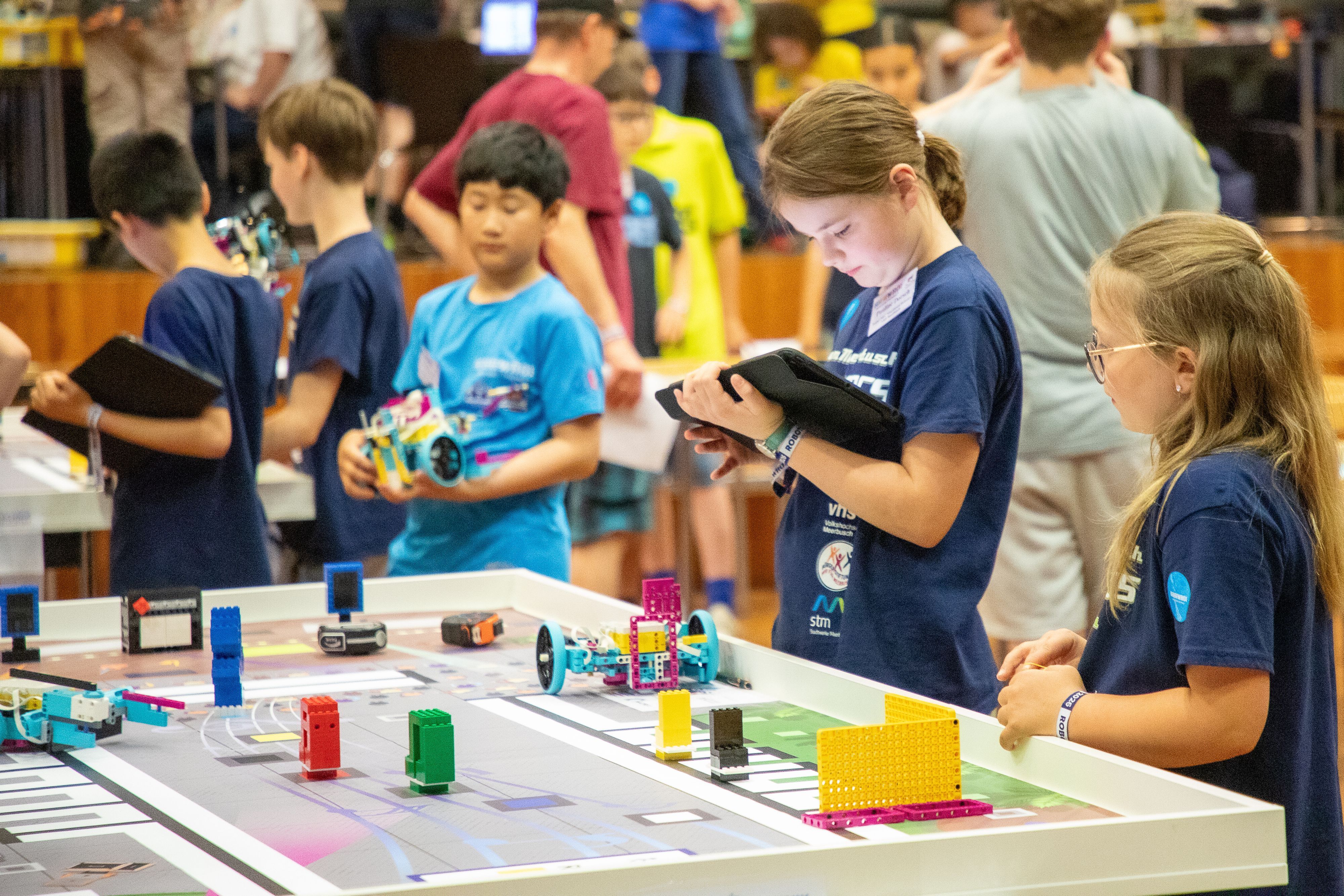 Kinder stehen Tischen mit zusammengesetzten Lego-Bauteilen, einige Kinder halten Tablets in den Händen.